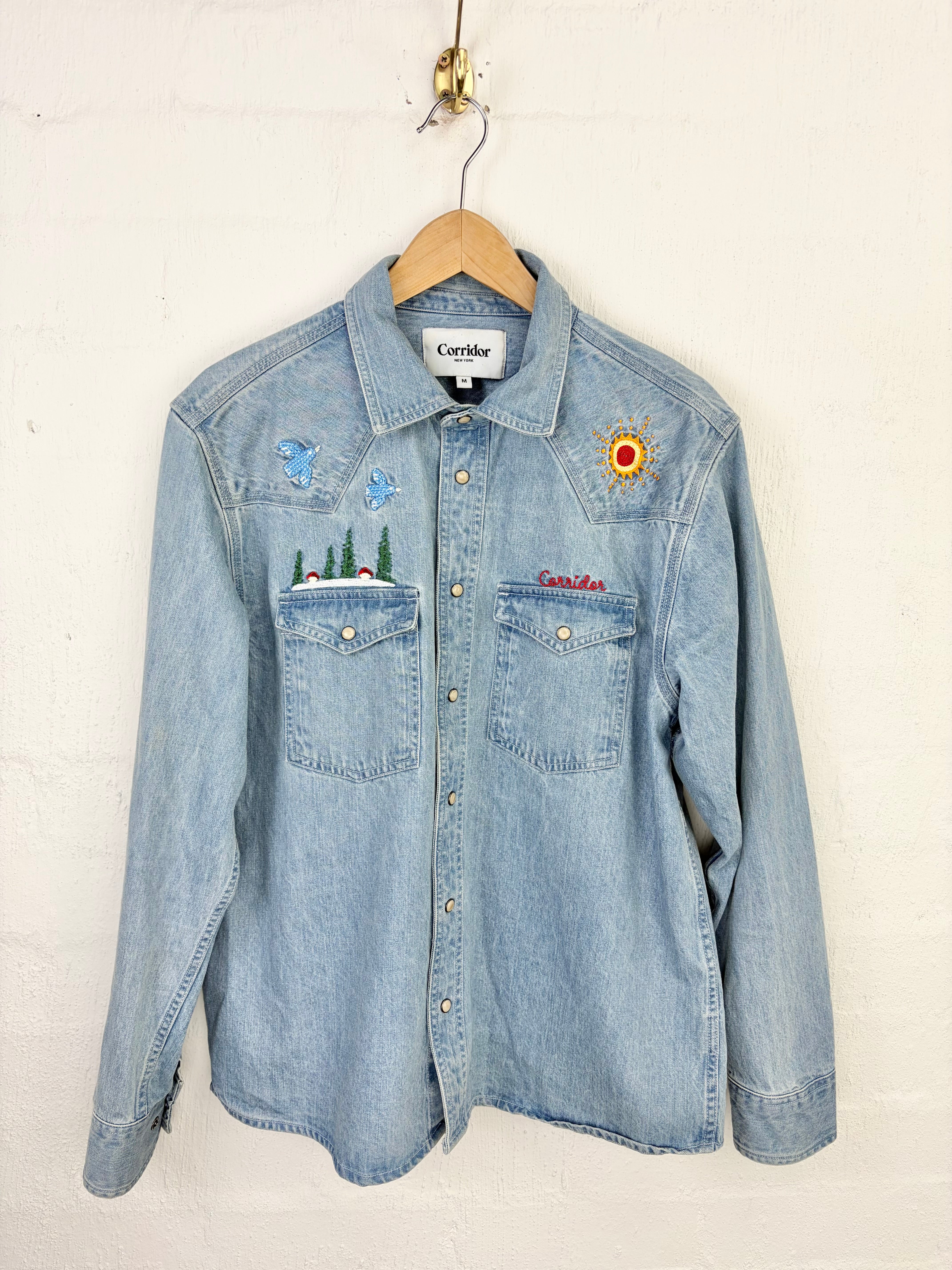A light blue denim western-style button-up shirt hanging on a wooden hanger against a white painted brick wall. The shirt features embroidered details, including a sun on the right chest, trees on the left chest pocket, and red script embroidery on the left chest pocket.
