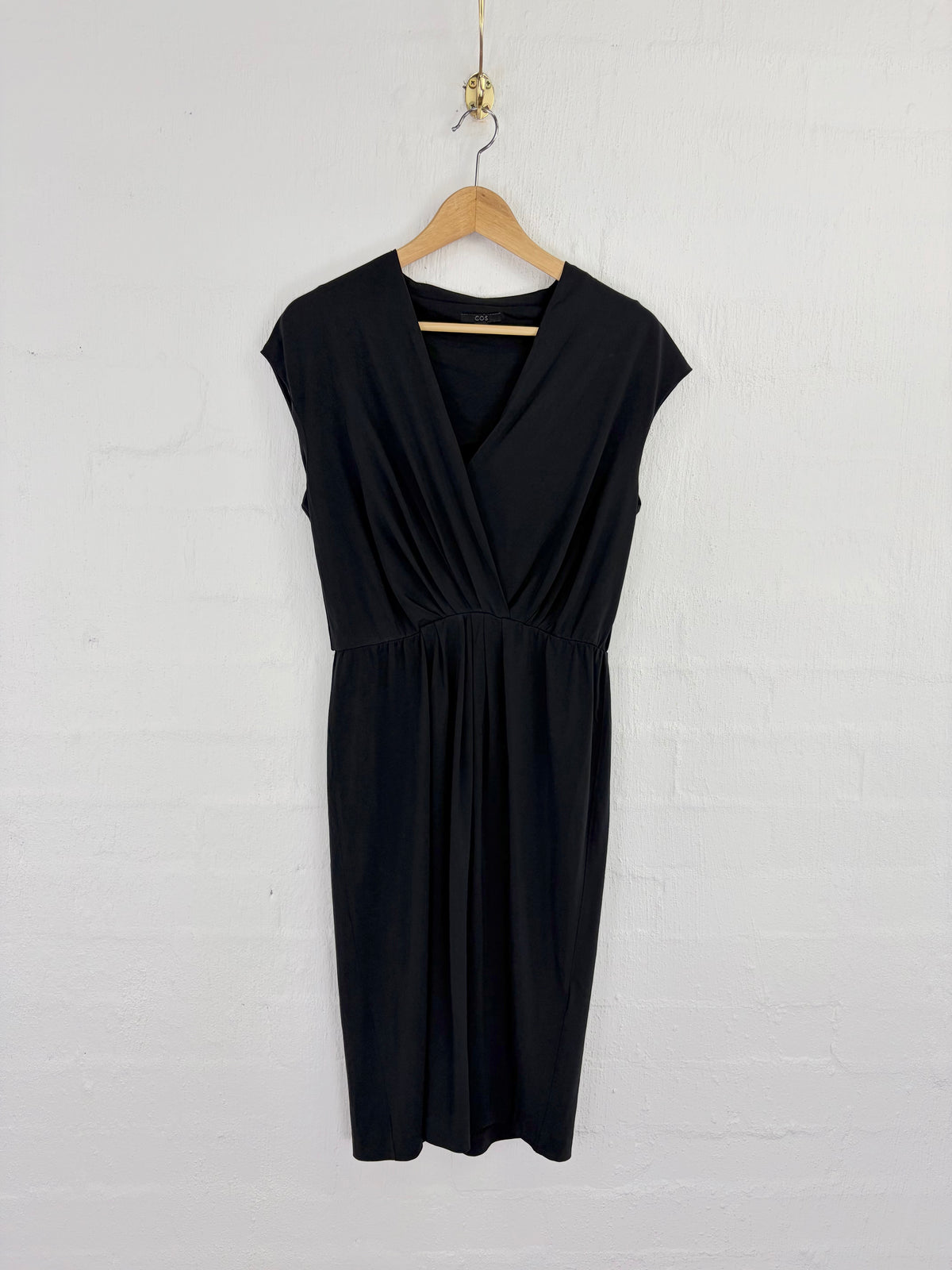 A black sleeveless wrap-style dress with a gathered waist, hanging on a wooden hanger against a white painted brick wall.