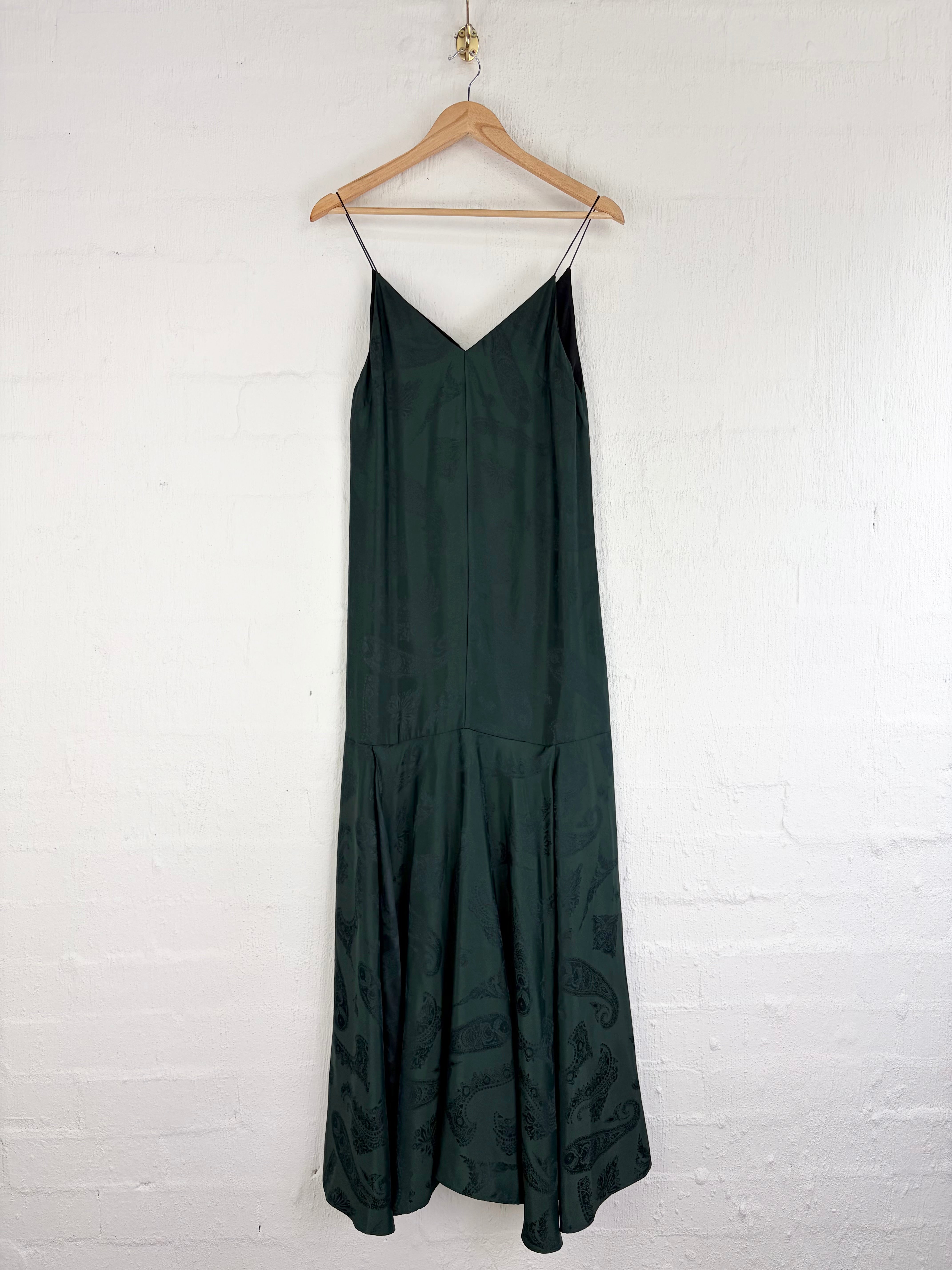 A dark forest green sleeveless slip dress with thin spaghetti straps and a V-neckline, hanging on a wooden hanger against a white painted brick wall.
