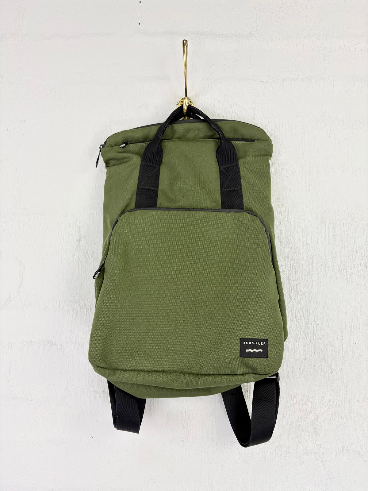 An olive green canvas backpack with black straps and a small black brand patch on the front, hanging from a gold hook against a white textured wall.