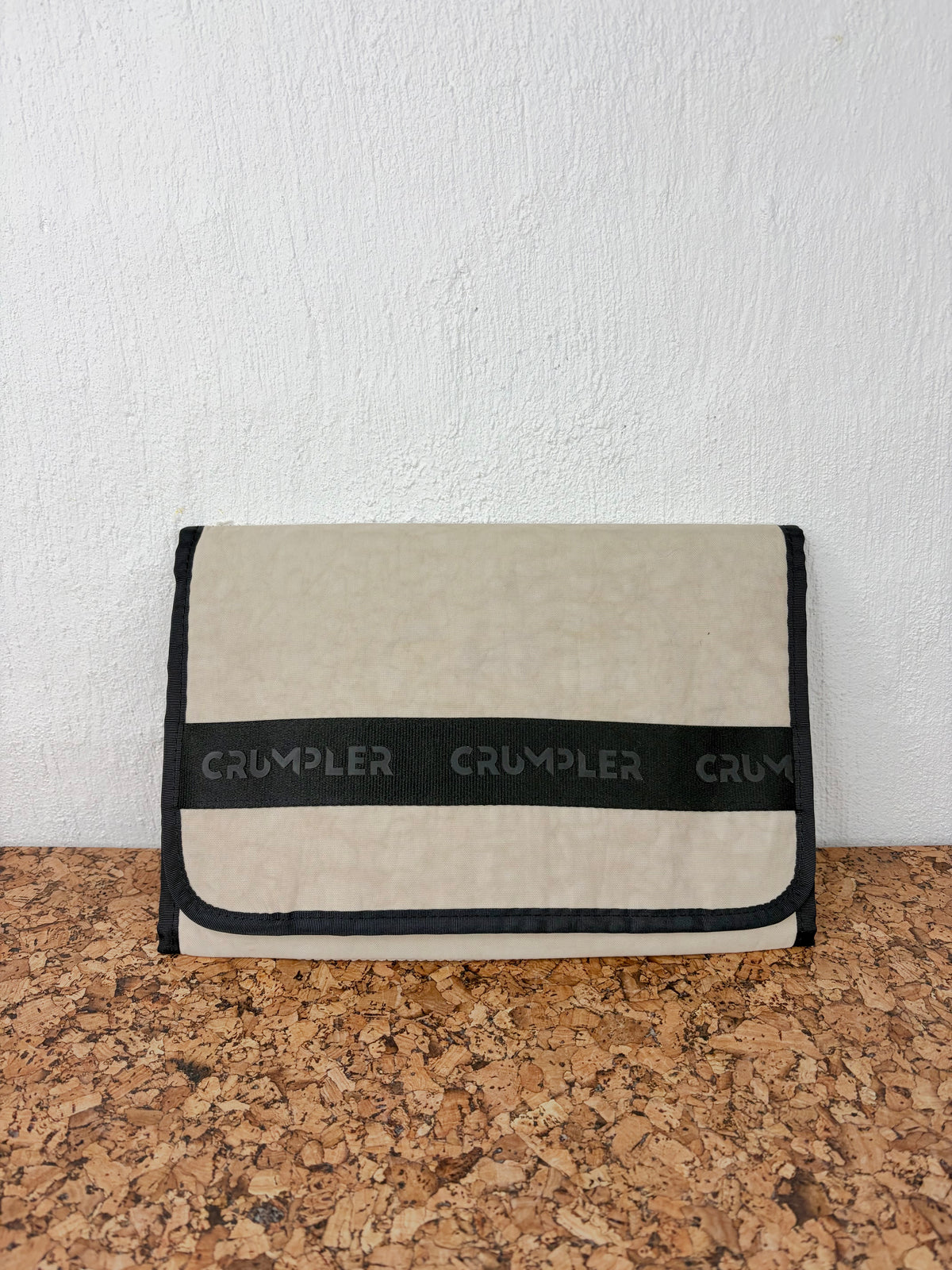 A beige textured fabric pouch with black trim and a black horizontal band featuring the brand name 'CRUMPLER' repeated, resting on a cork surface against a white wall.