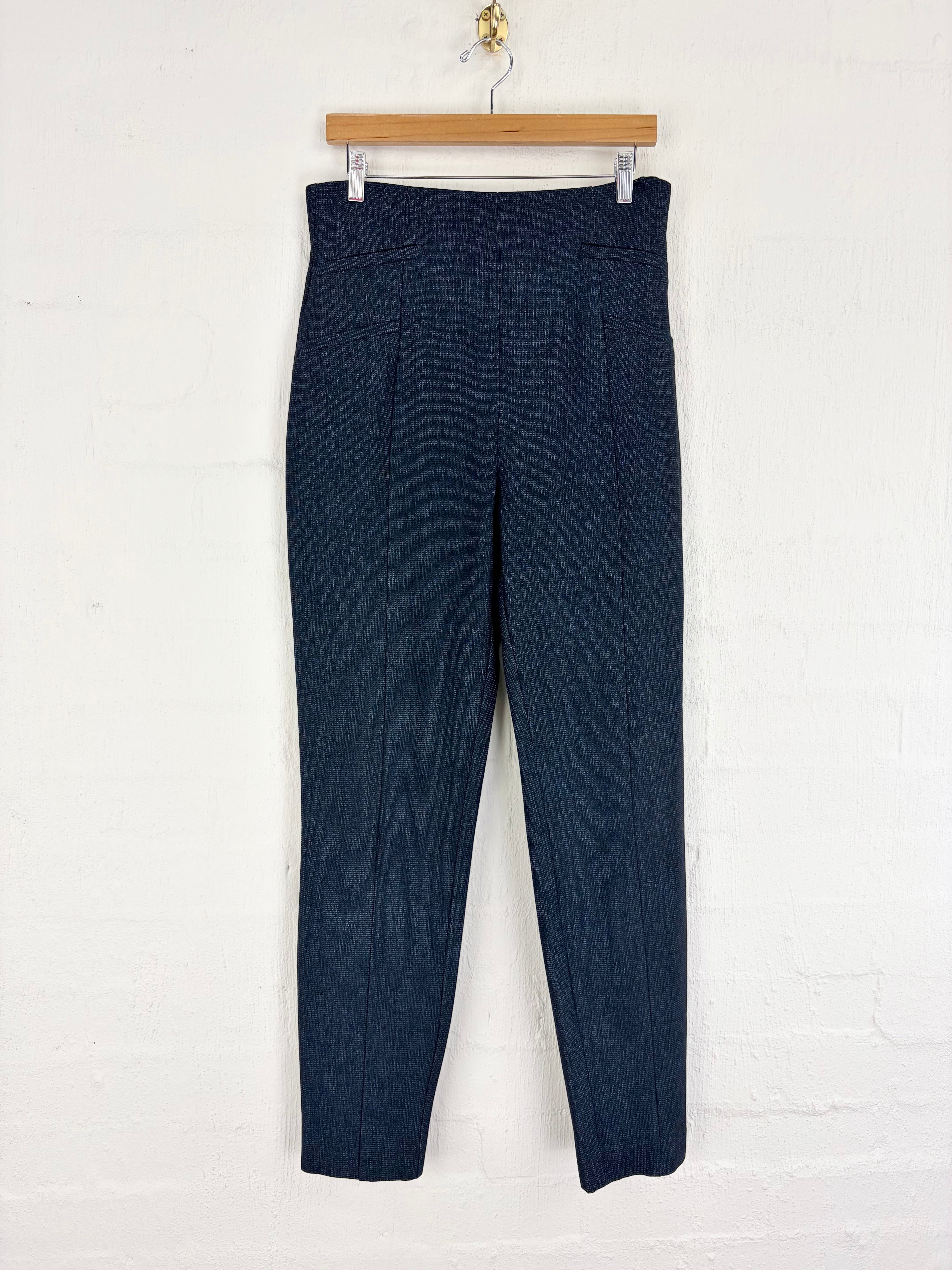 Dark navy blue trousers hanging from a wooden hanger against a white painted brick wall.