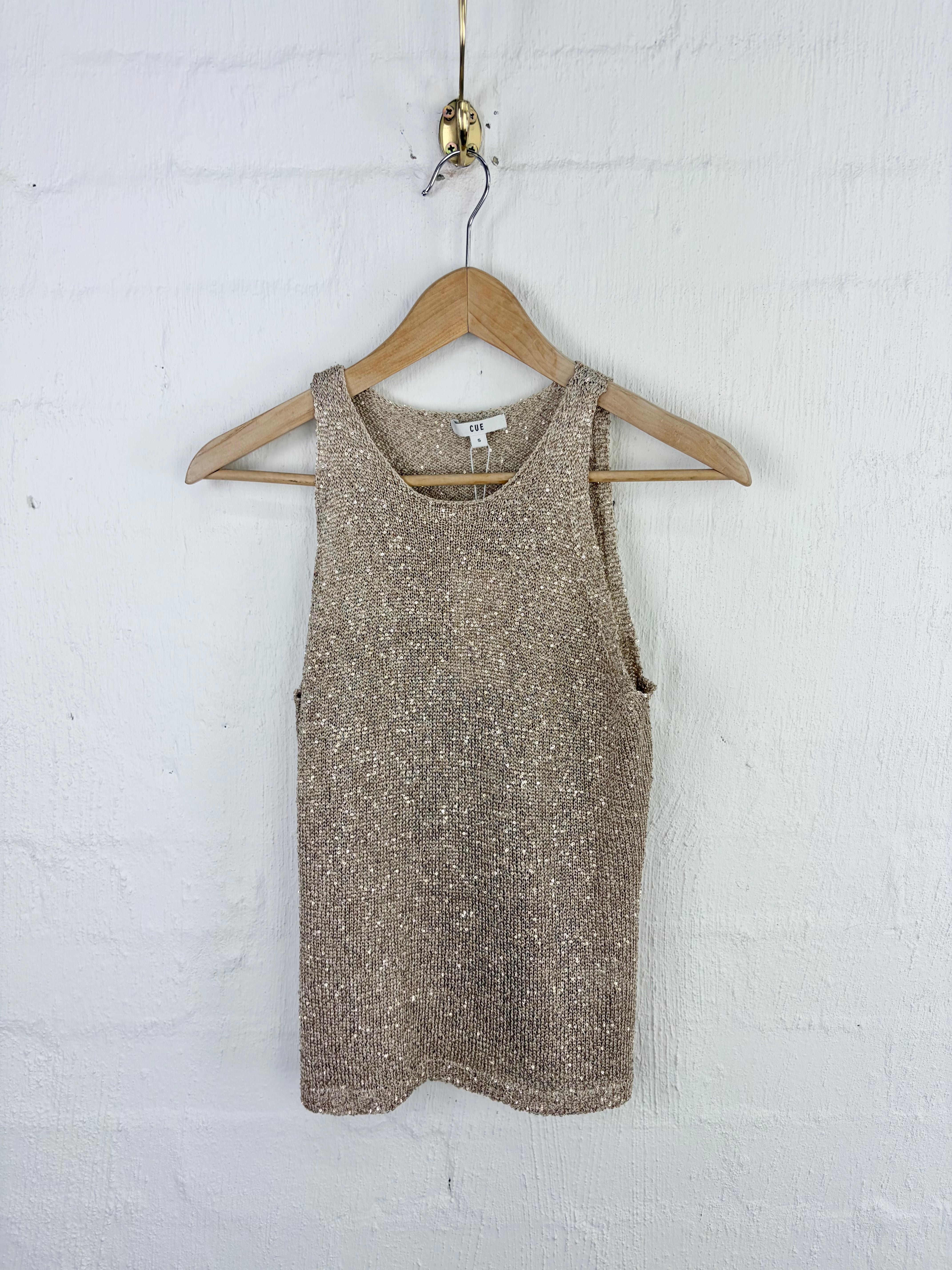 A sleeveless knit top with a textured, sparkly metallic finish in a champagne-gold tone, hanging on a wooden hanger against a white painted brick wall.