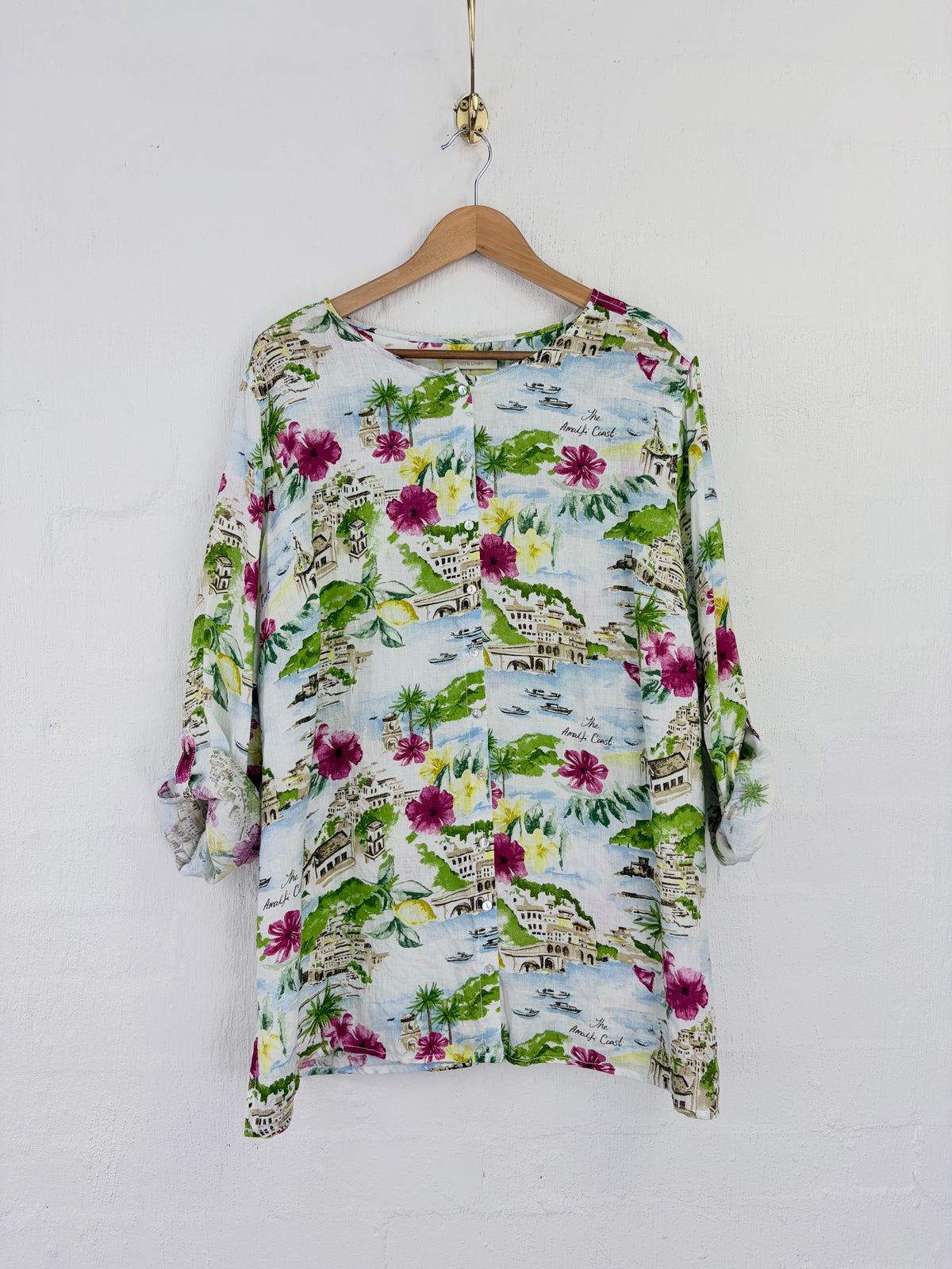 A long-sleeve button-up blouse with a colorful tropical landscape print, hanging on a wooden hanger against a white painted brick wall.