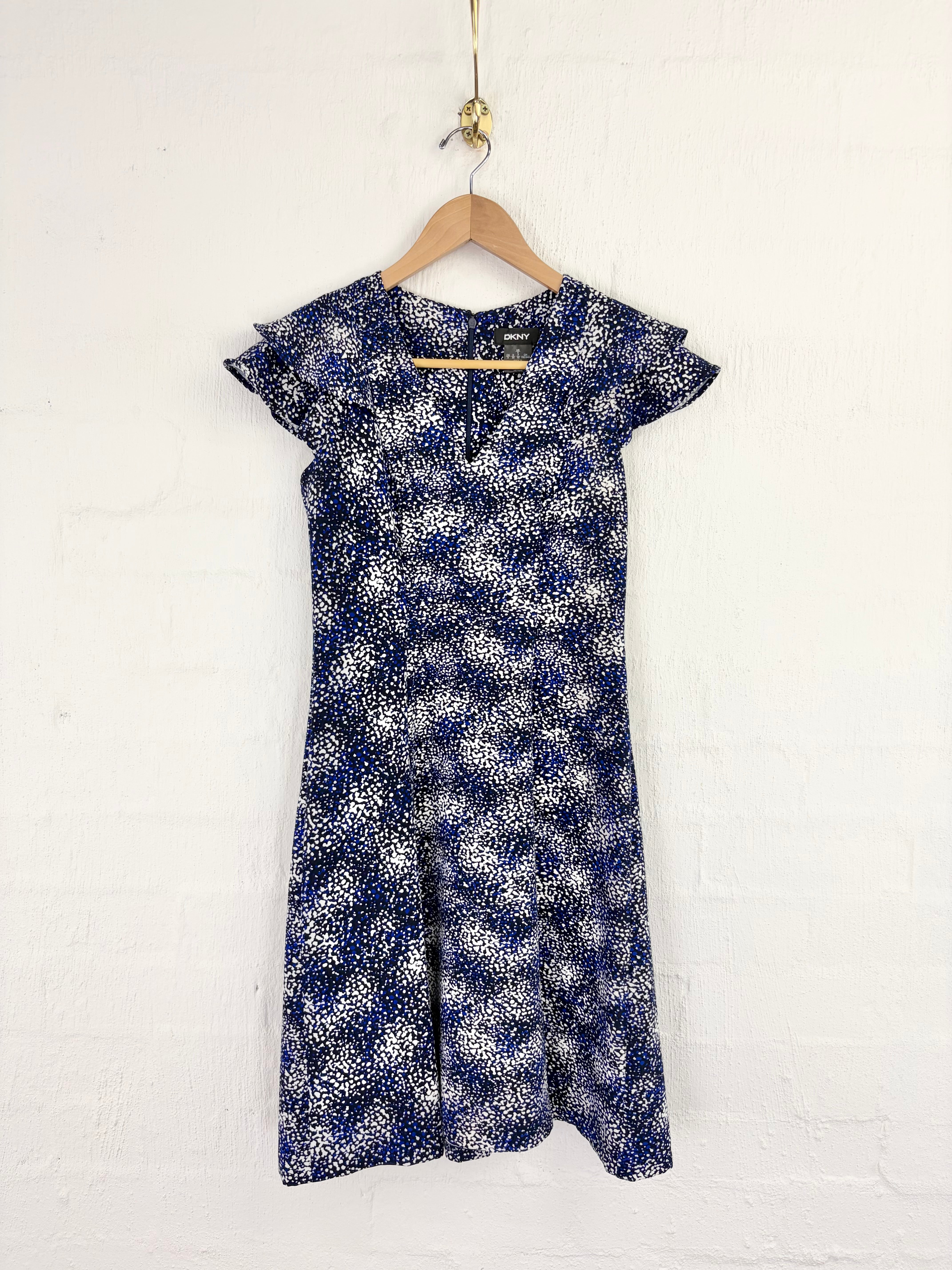 A short-sleeve knee-length dress with a blue and white abstract floral pattern, hanging on a wooden hanger against a white painted brick wall.