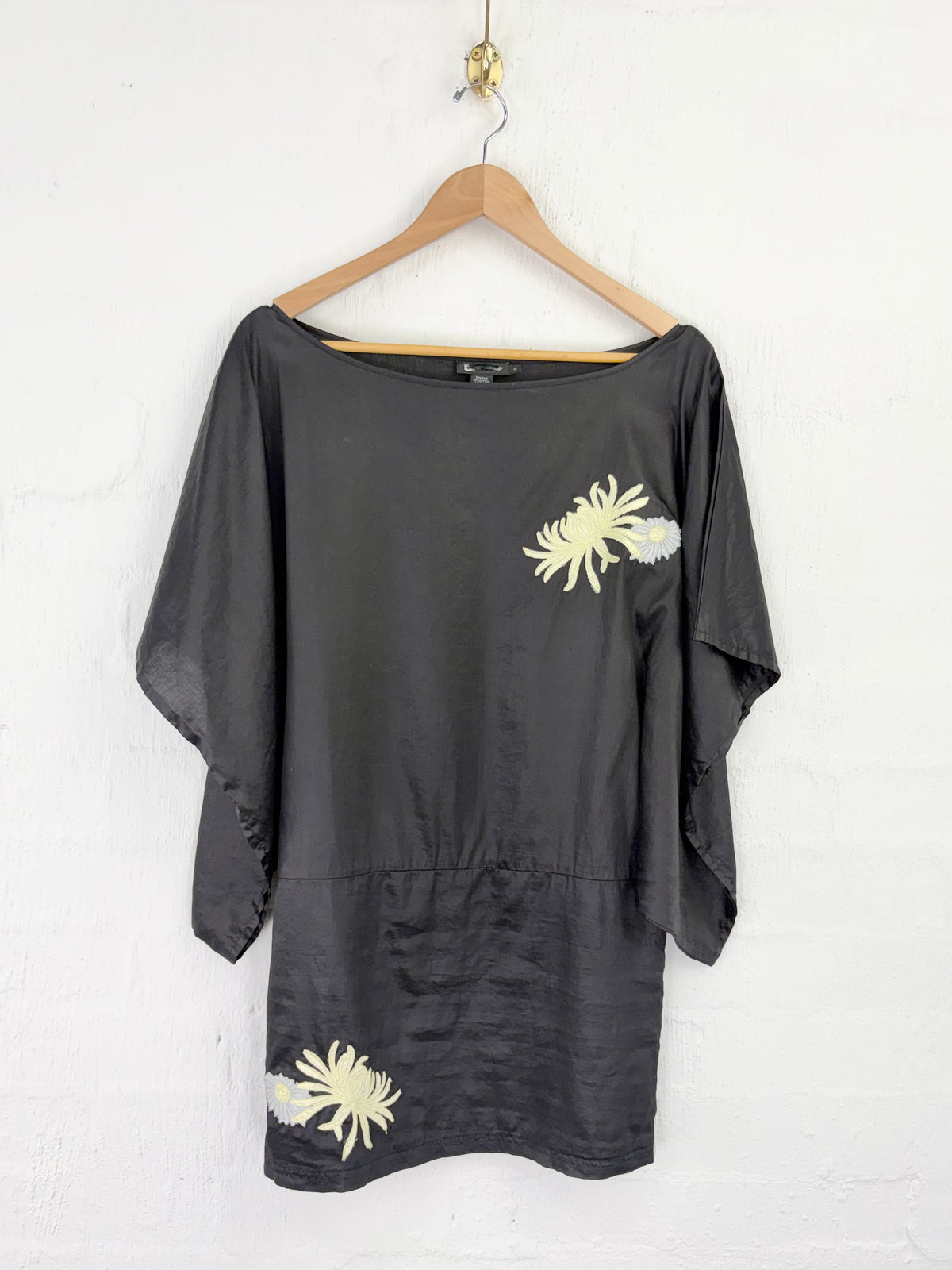 A black wide-neck tunic top with three-quarter sleeves, featuring two cream-colored floral embroidery details, hanging on a wooden hanger against a white painted brick wall.