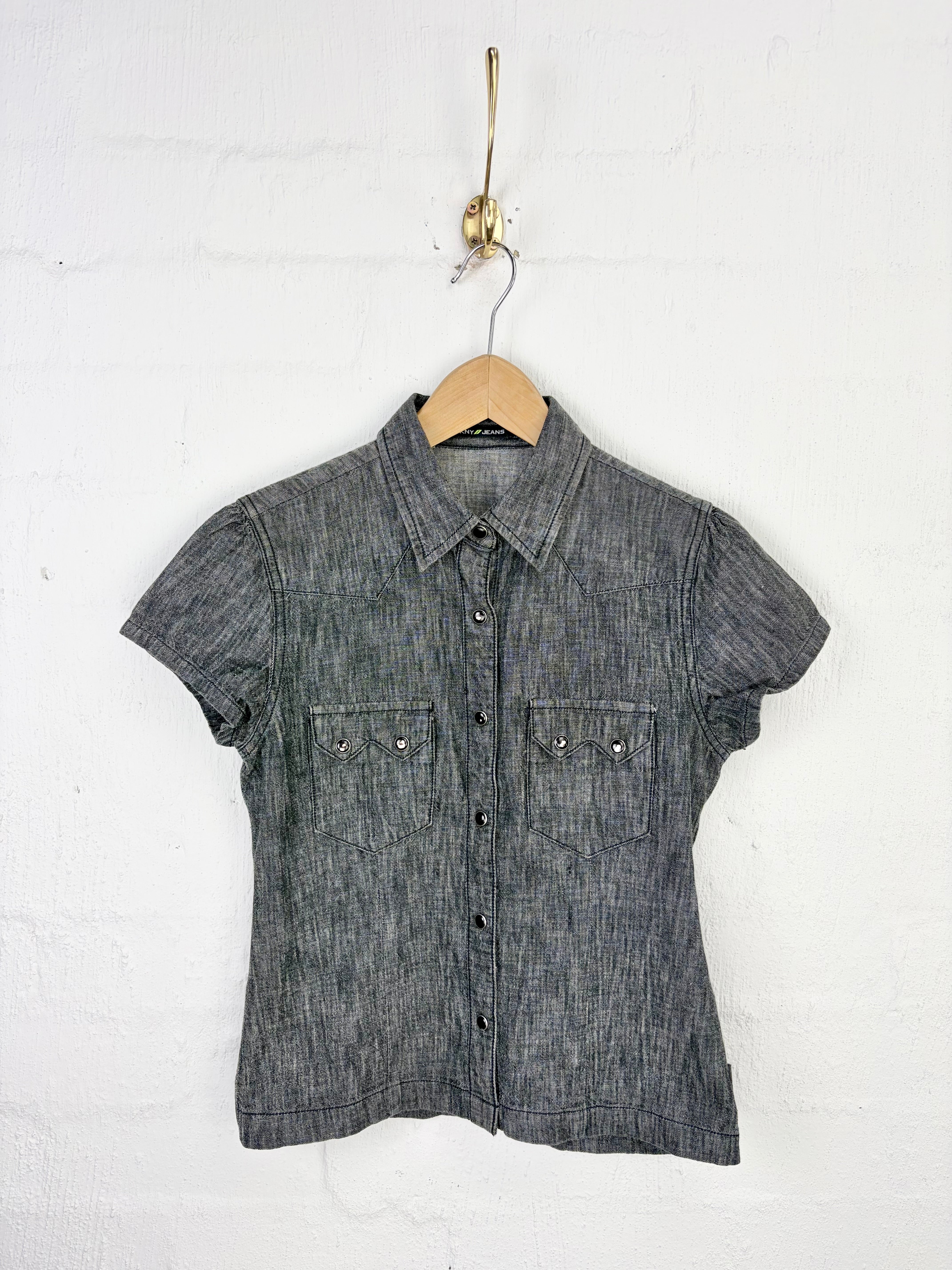 A short-sleeve button-up shirt in a dark gray textured chambray fabric, hanging on a wooden hanger from a gold hook against a white painted brick wall.
