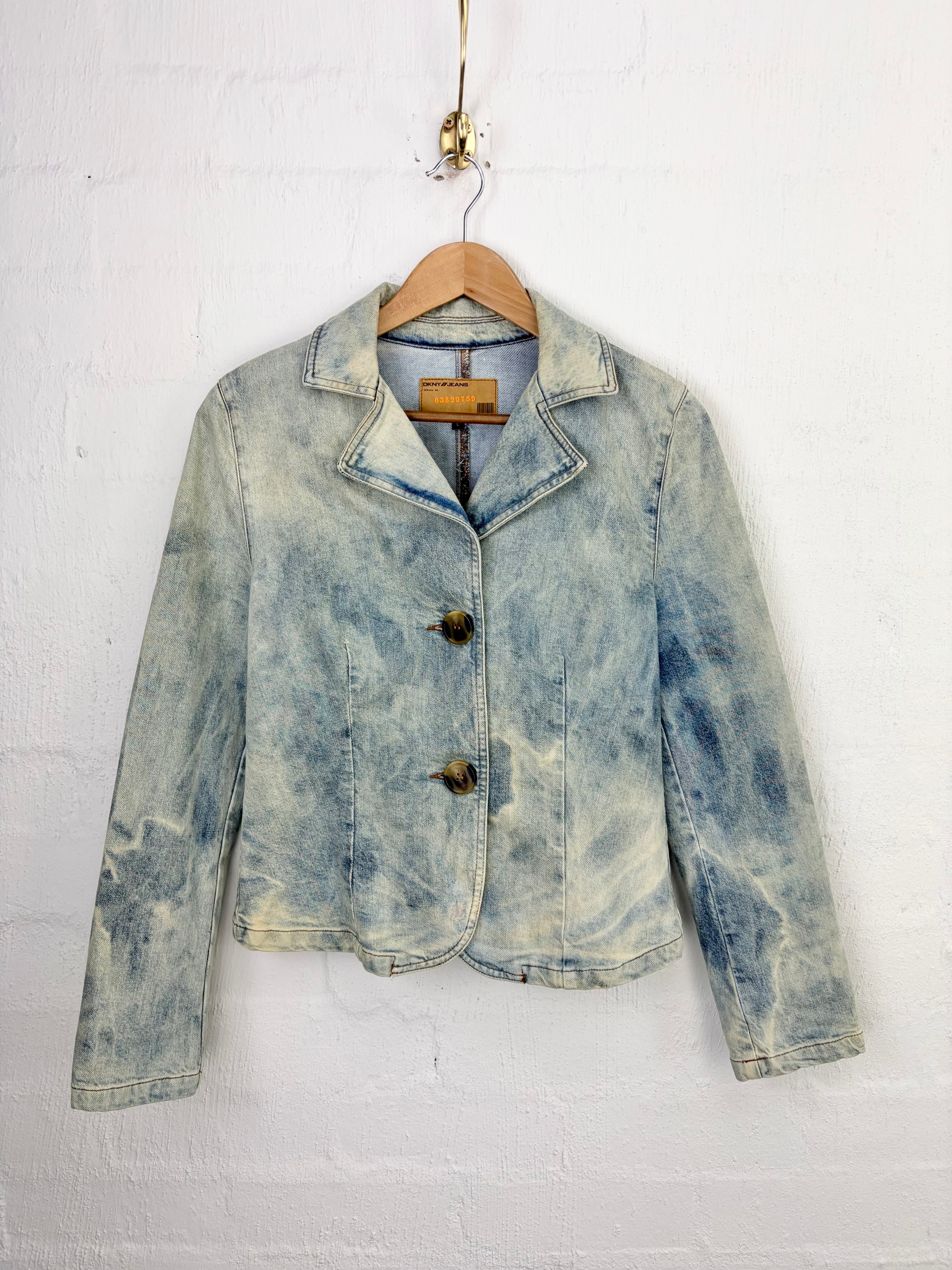 A cropped, light-wash blue denim jacket with a mineral or acid-wash finish, hanging on a wooden hanger against a white painted brick wall.