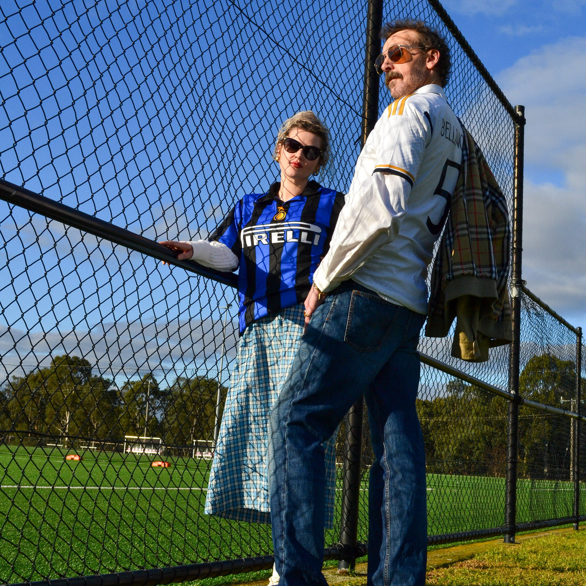 Two people standing outdoors beside a chain-link fence on a grassy field under a blue sky. One person wears a blue and black striped jersey with 'MELBOURNE' across the chest, and the other wears a white long-sleeve shirt and jeans.