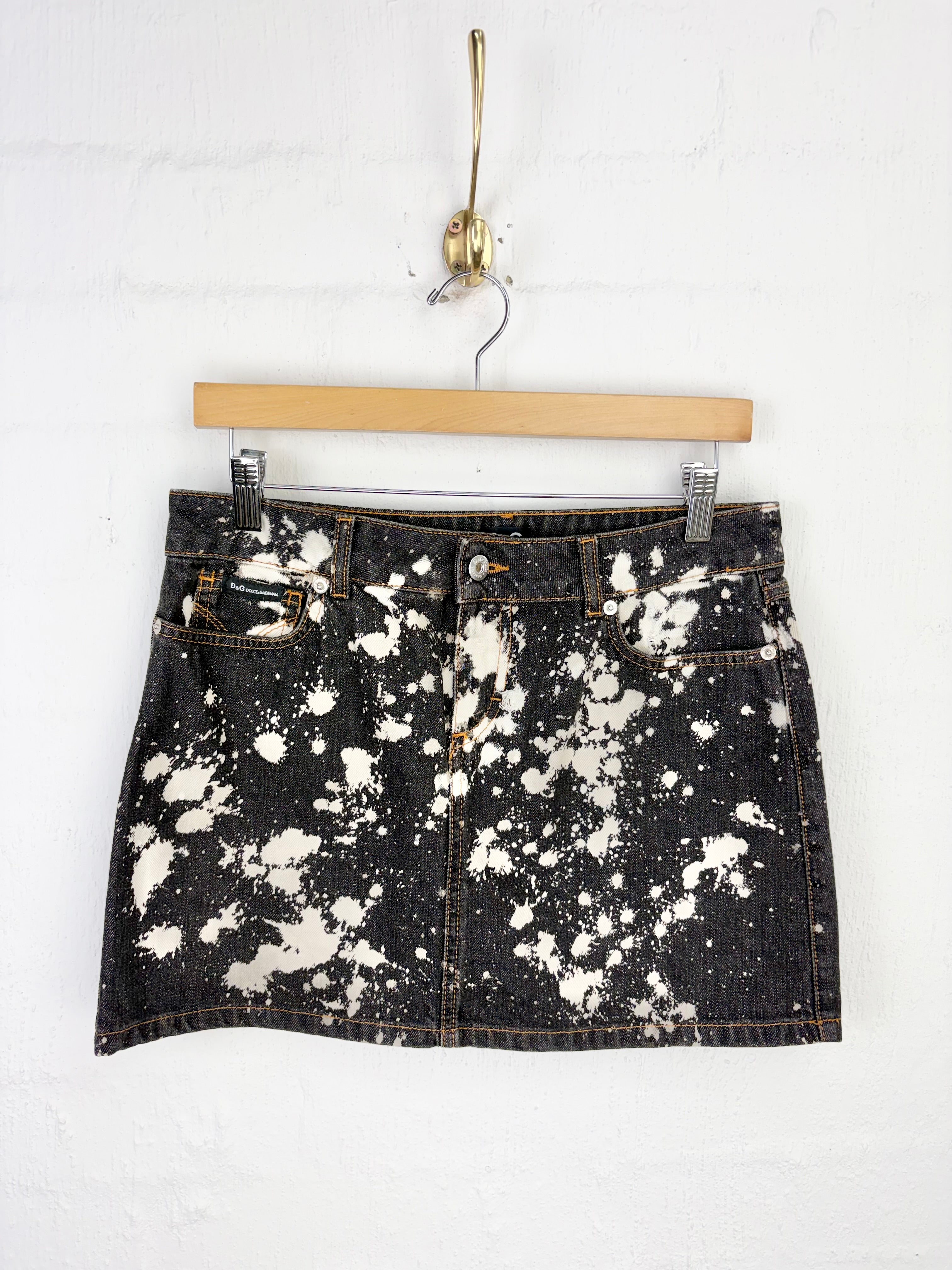 A black denim mini skirt with a white bleach-splatter or paint-splatter pattern, hanging from a wooden hanger on a white brick wall.
