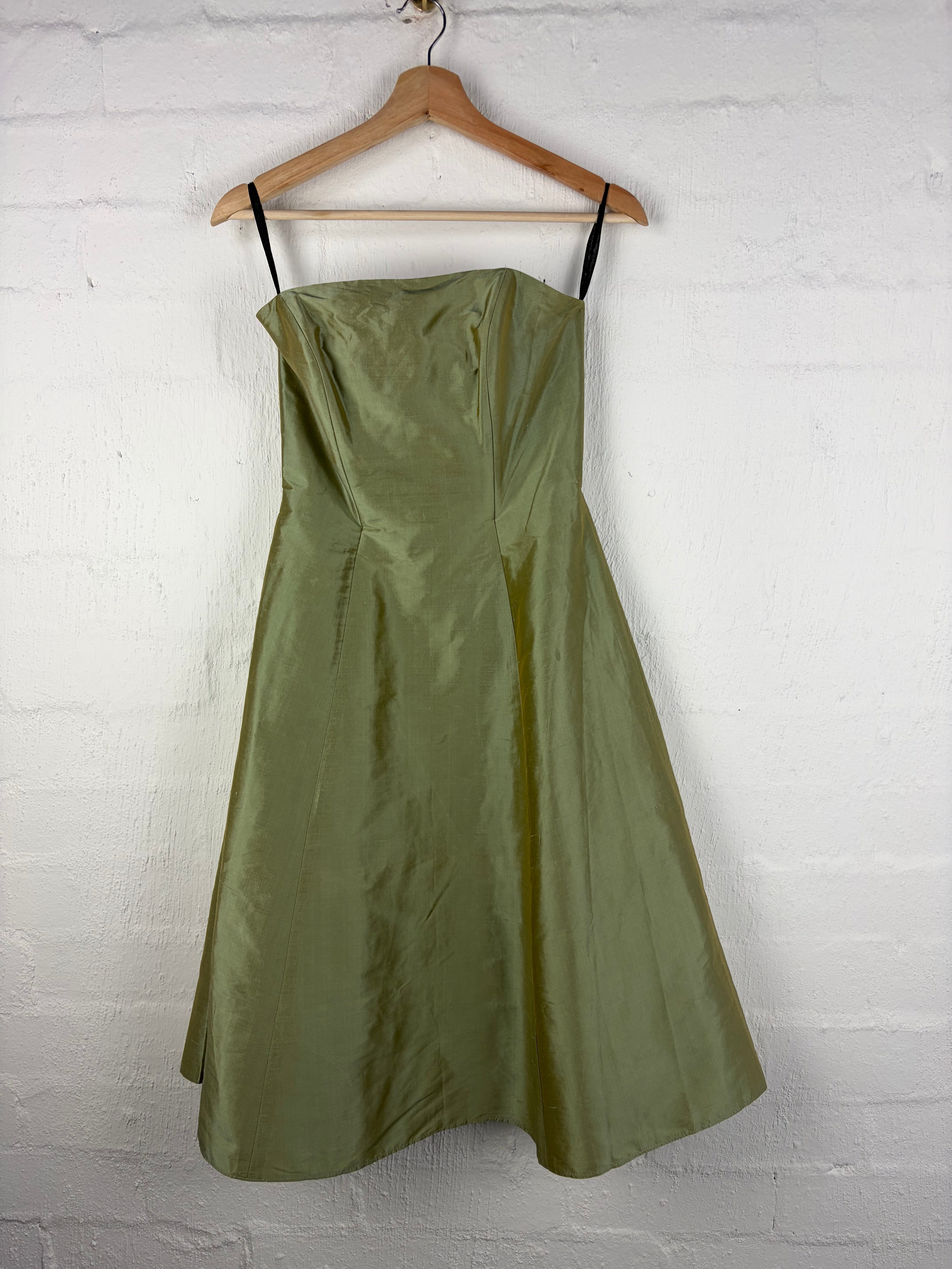An olive green satin slip dress draped over a wooden hanger against a white painted brick wall.