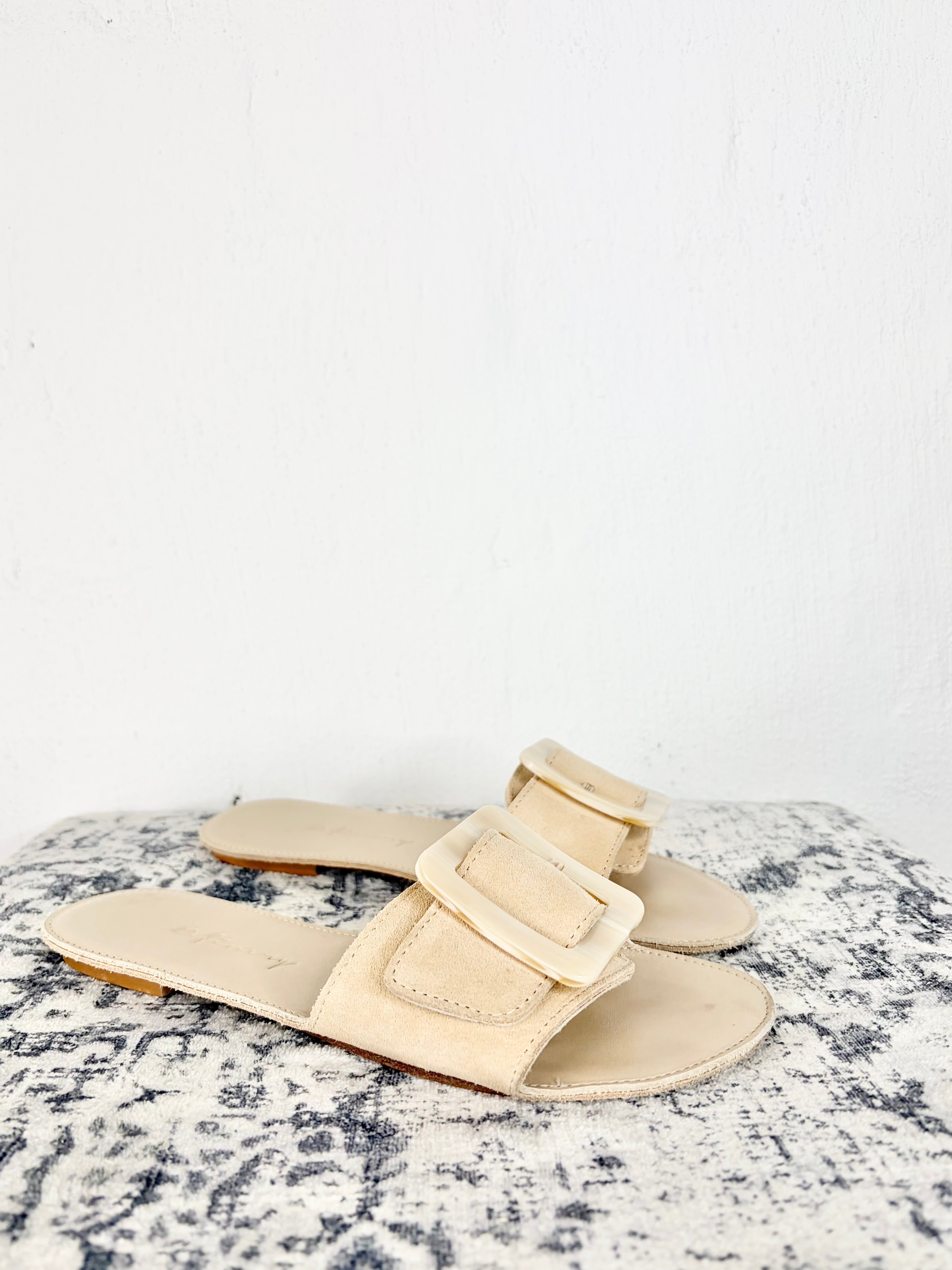 A pair of beige double-strap slide sandals with large square buckle details, displayed on a textured black and white surface against a plain white wall.