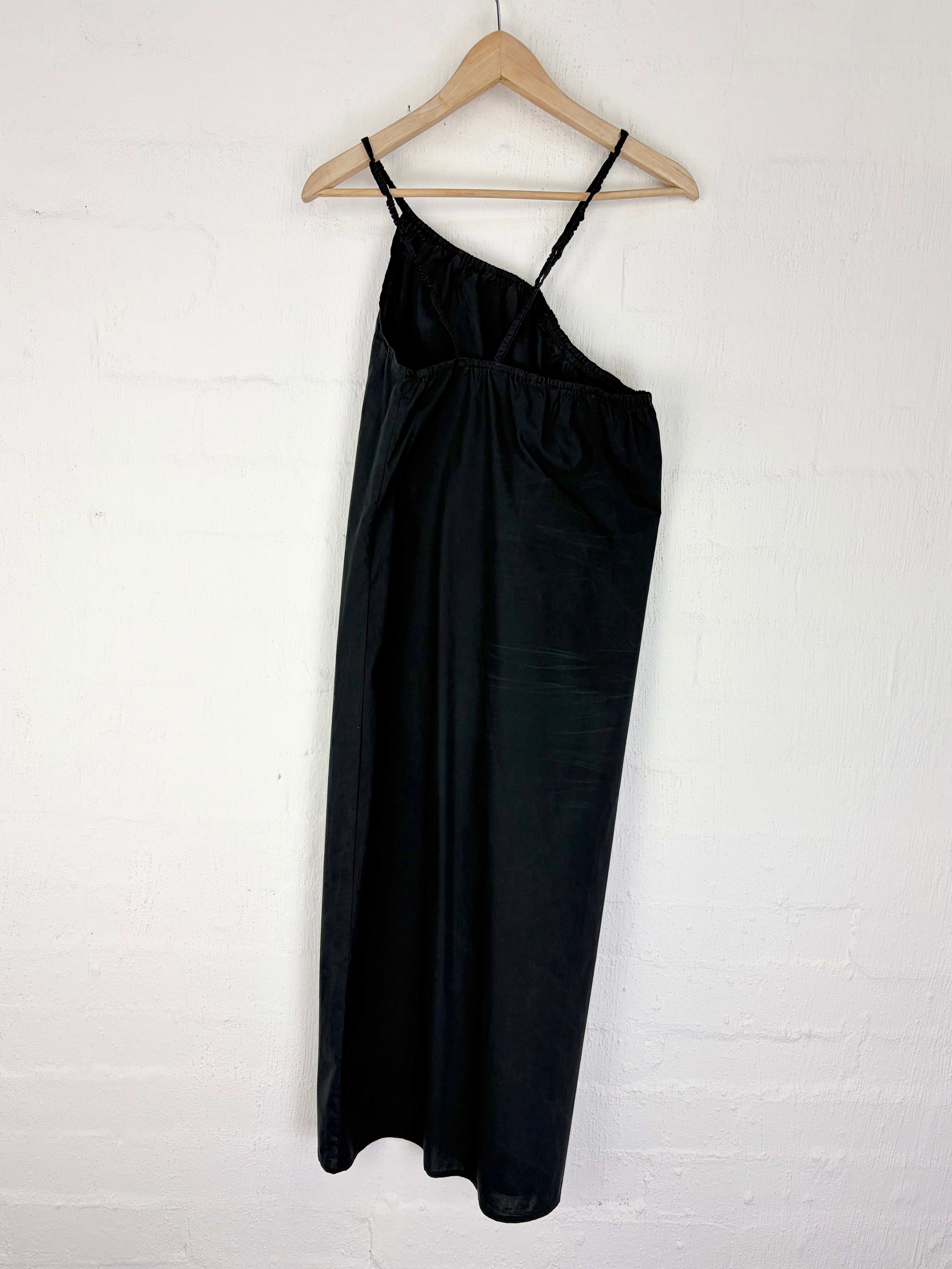 A black satin slip dress with a single shoulder strap, hanging on a wooden hanger against a white painted brick wall.