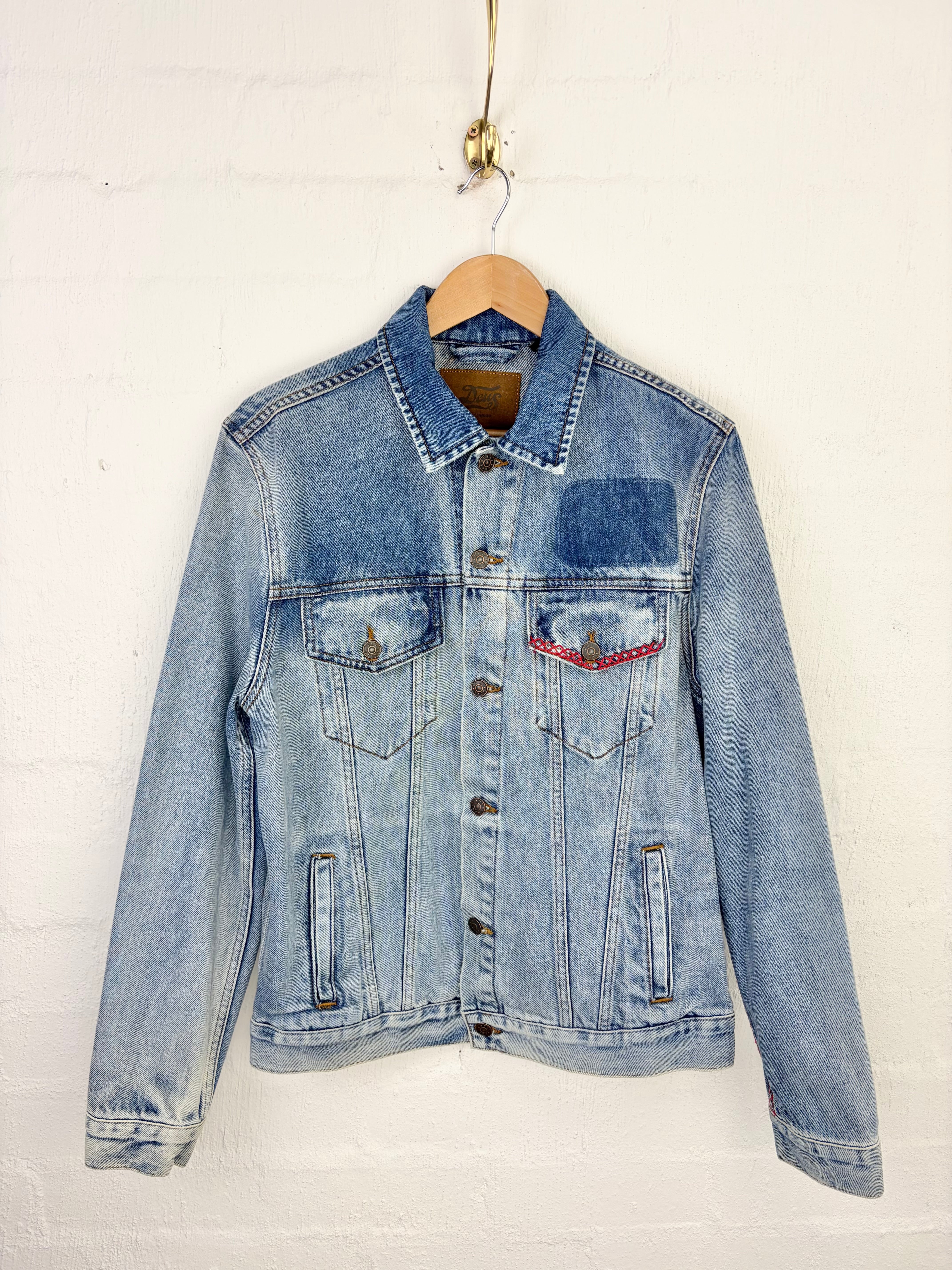 A light blue denim jacket with a patch pocket and a red embroidered detail on the chest pocket, hanging on a wooden hanger against a white painted brick wall.