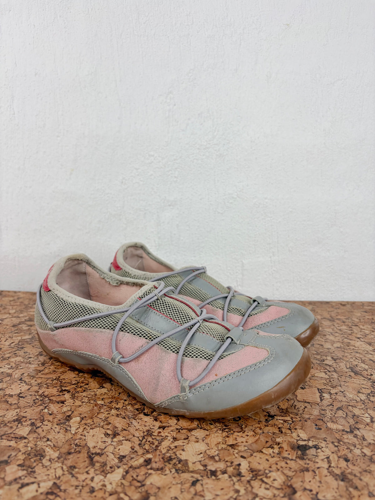 A pair of children's slip-on shoes with a gray and pink color-blocked design, featuring gray laces and a brown rubber sole, resting on a cork surface against a white wall.