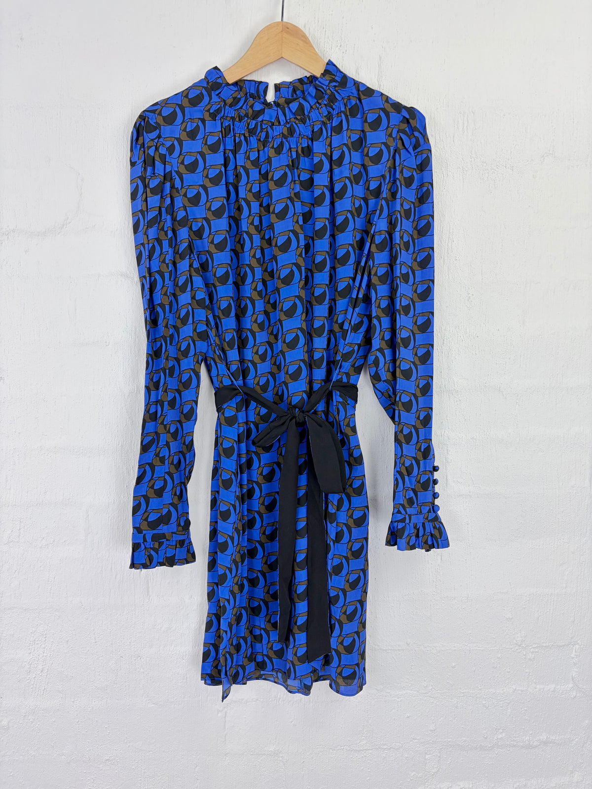 A long-sleeve blue and black patterned dress with a high gathered neckline and a black waist tie, hanging on a wooden hanger against a white brick wall.