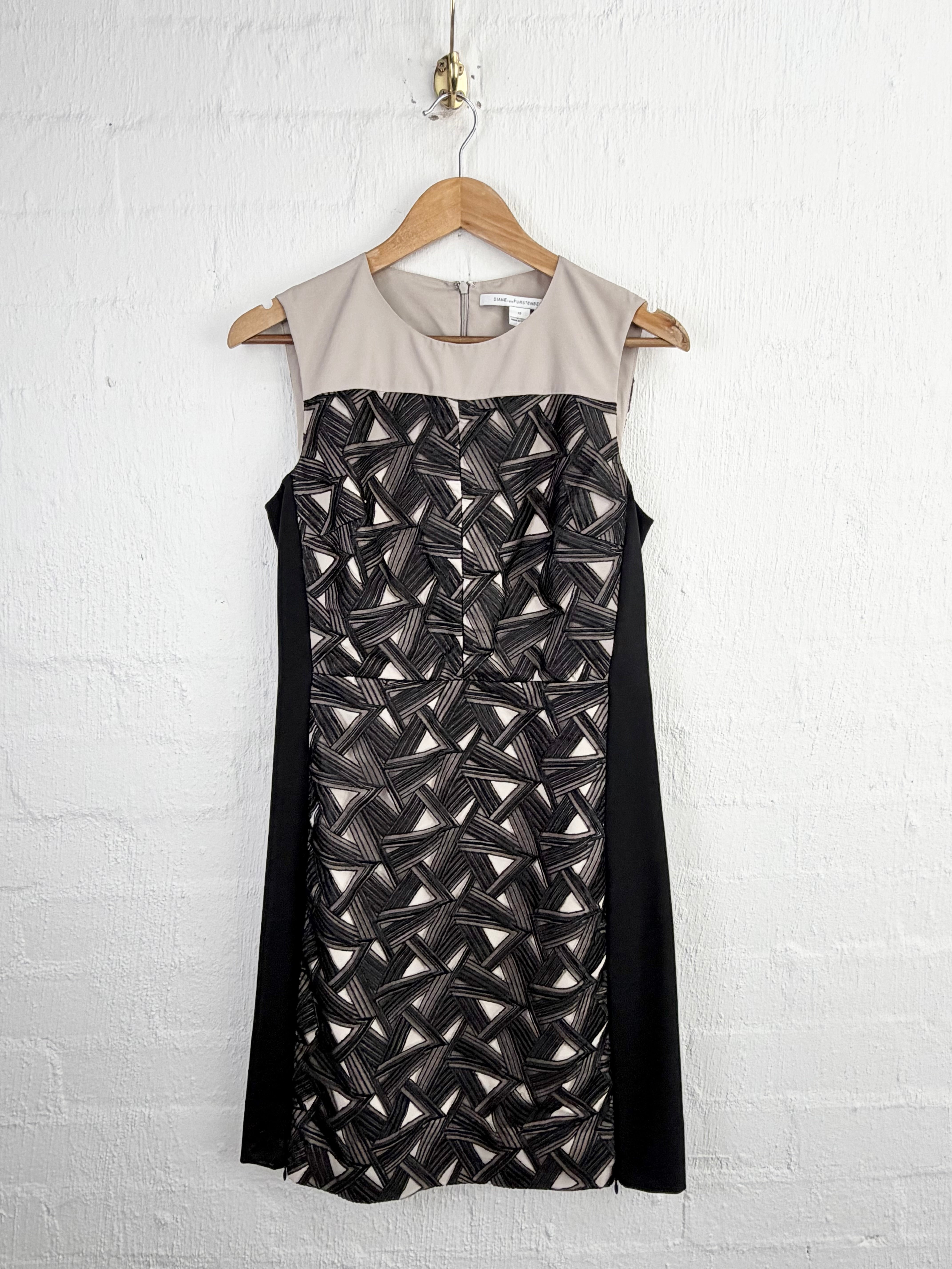 A sleeveless sheath dress hanging on a wooden hanger against a white painted brick wall. The dress has a light beige crew neck and upper yoke, with black side panels and a geometric patterned center panel in black, gray, and metallic silver.