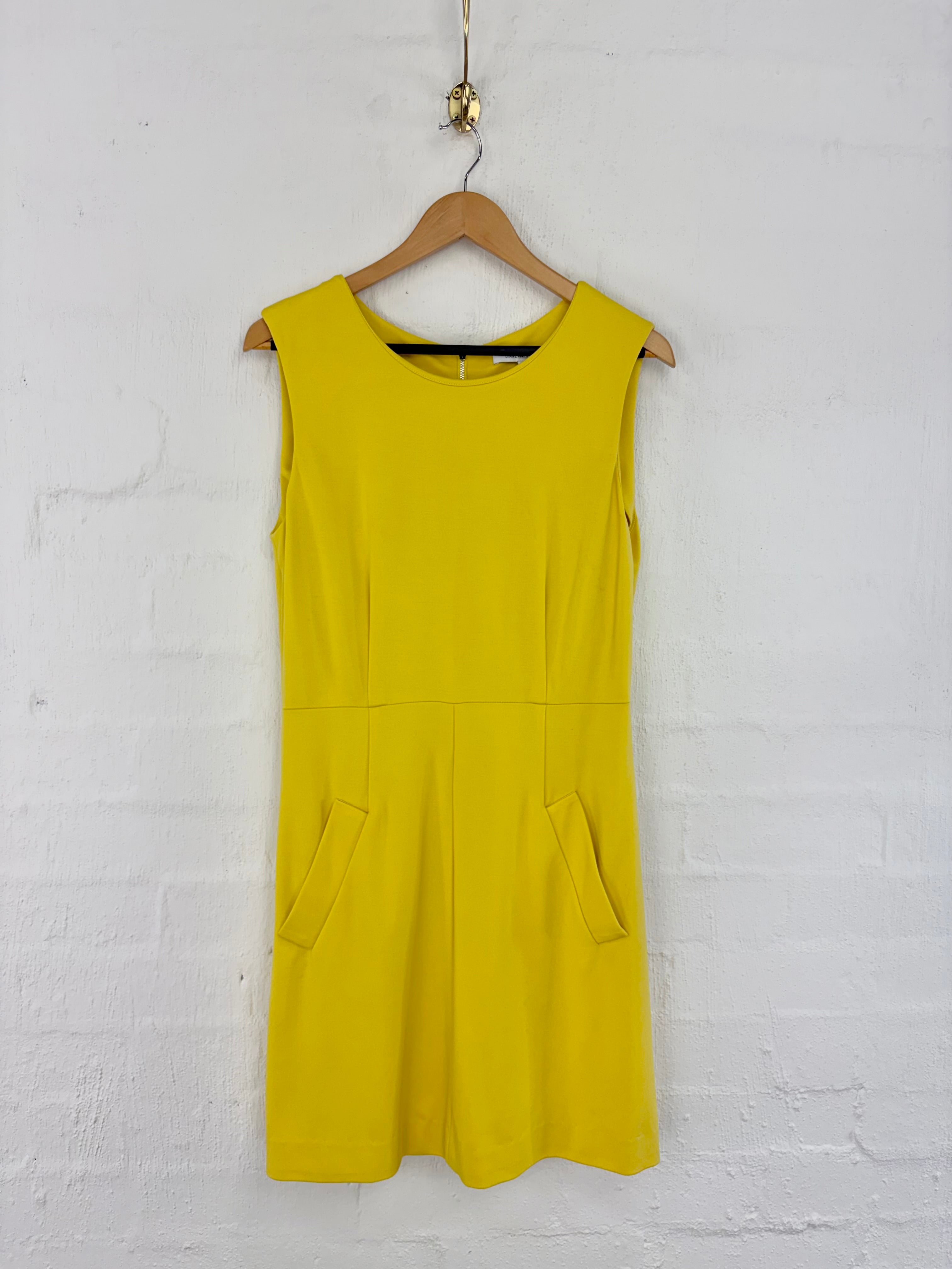 A bright yellow sleeveless shift dress with two front patch pockets, hanging on a wooden hanger against a white painted brick wall.