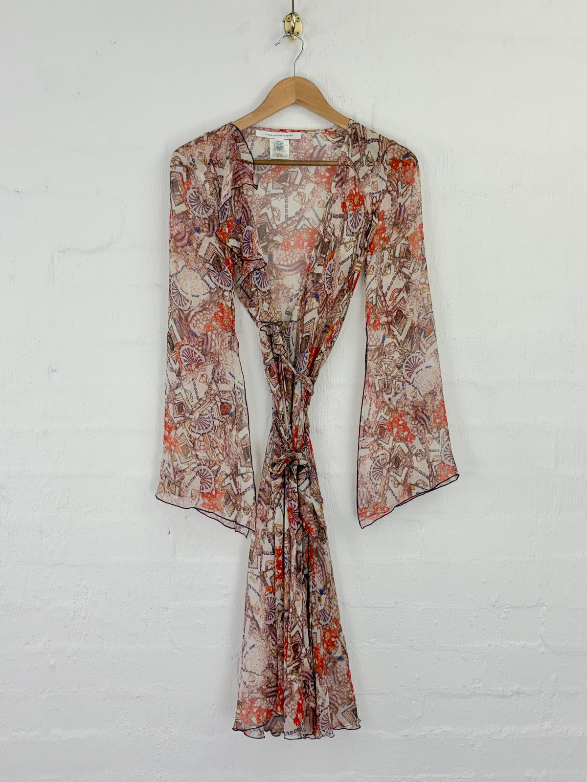 A long-sleeve wrap-style dress with a tie waist, featuring an abstract floral and paisley print in rust orange, brown, and beige tones, hanging on a wooden hanger against a white brick wall.