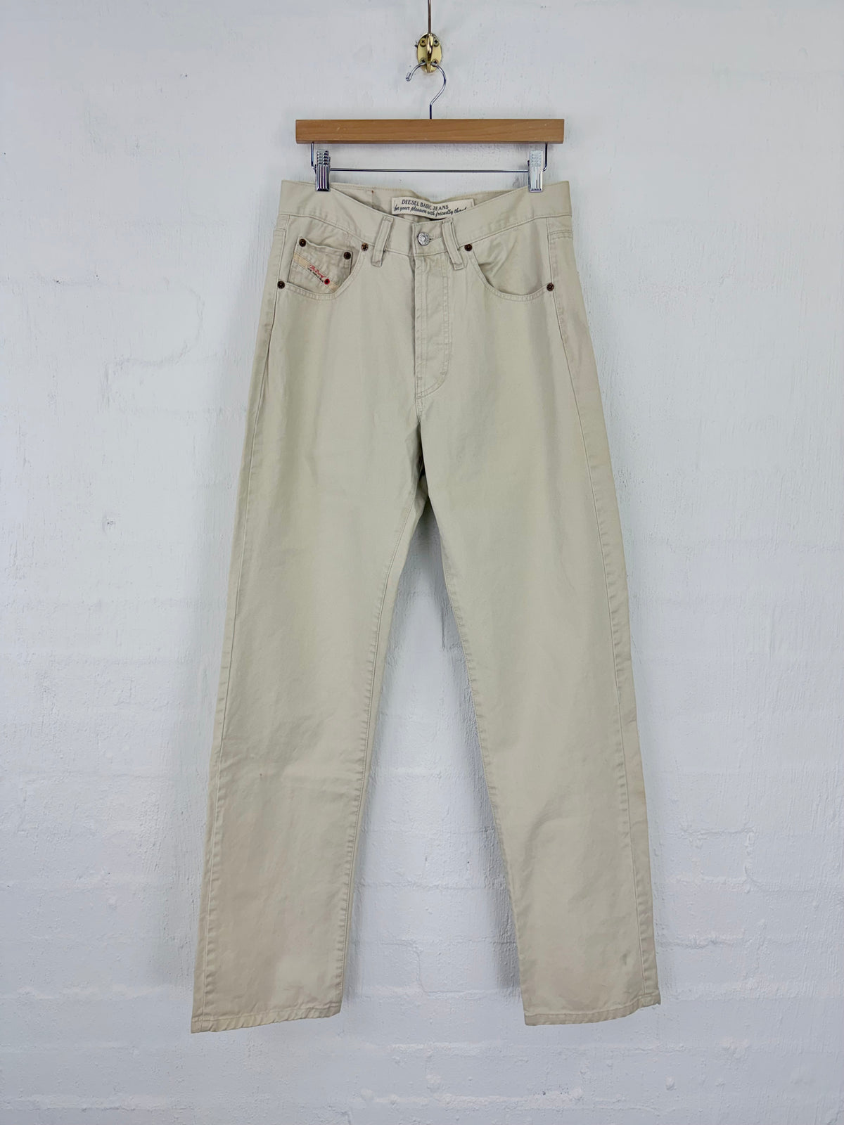 A pair of light beige straight-leg jeans hanging from a wooden hanger against a white painted brick wall.