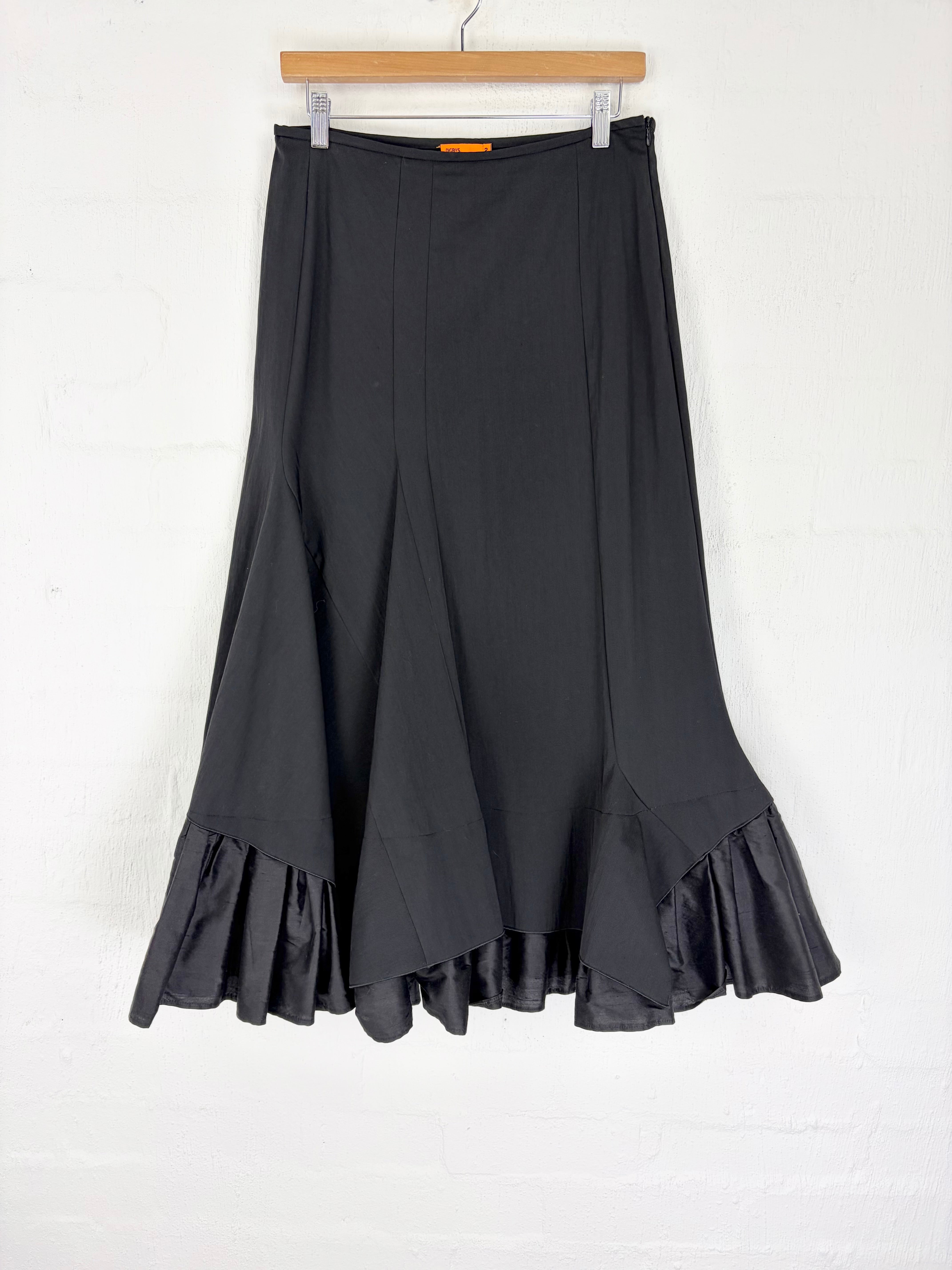 A black midi skirt with a flared, ruffled hem, hanging from a wooden hanger against a white painted brick wall.
