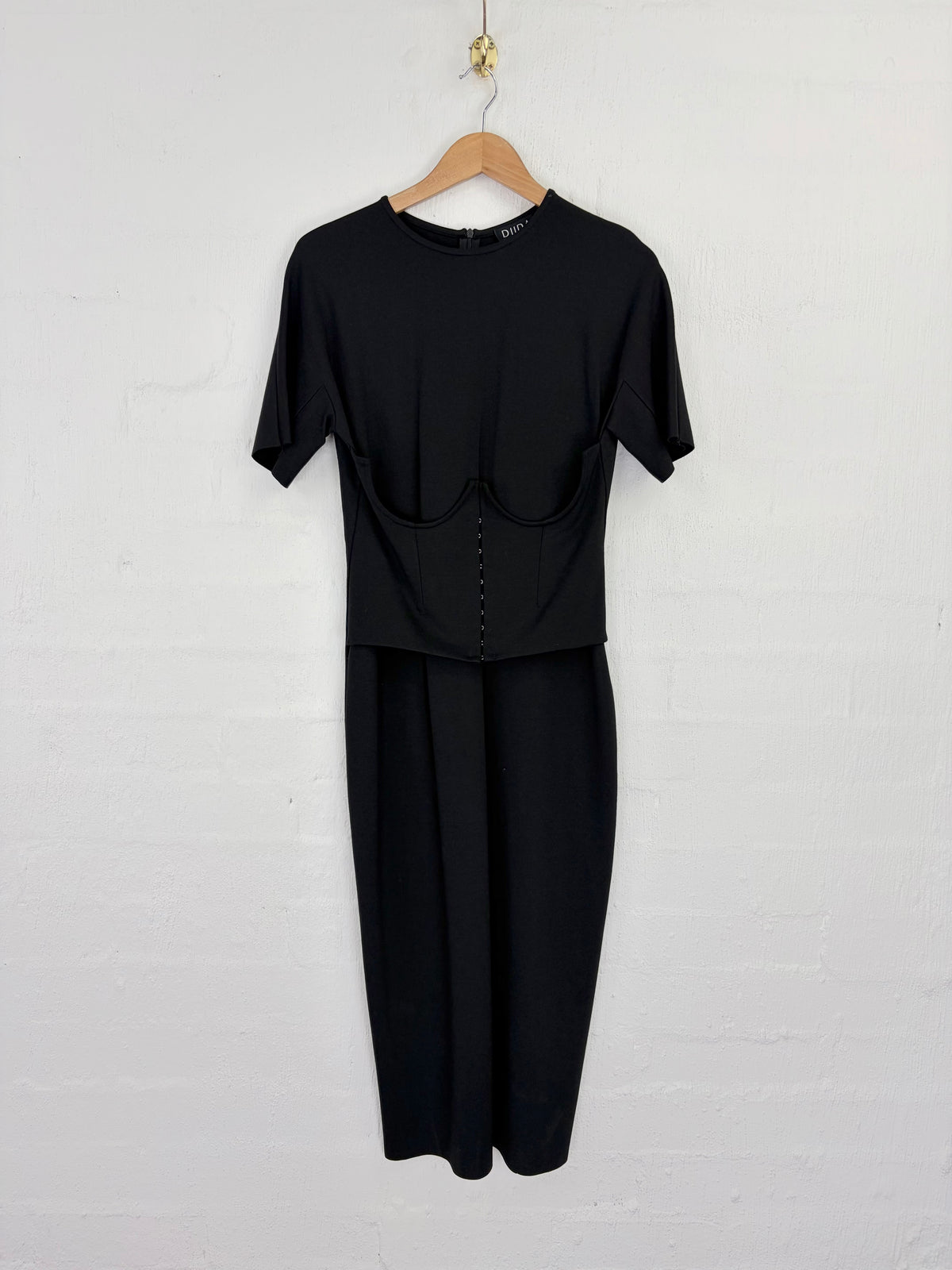 A black short-sleeve dress with a structured corset-style bodice and matching wide-leg skirt, hanging on a wooden hanger against a white painted brick wall.