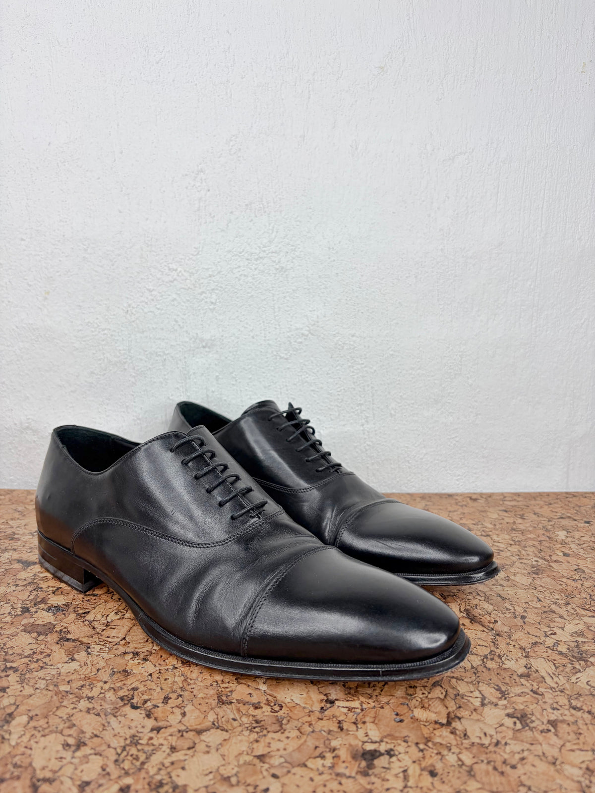 A pair of black leather cap-toe oxford shoes with black laces, displayed on a brown cork surface against a white textured wall.