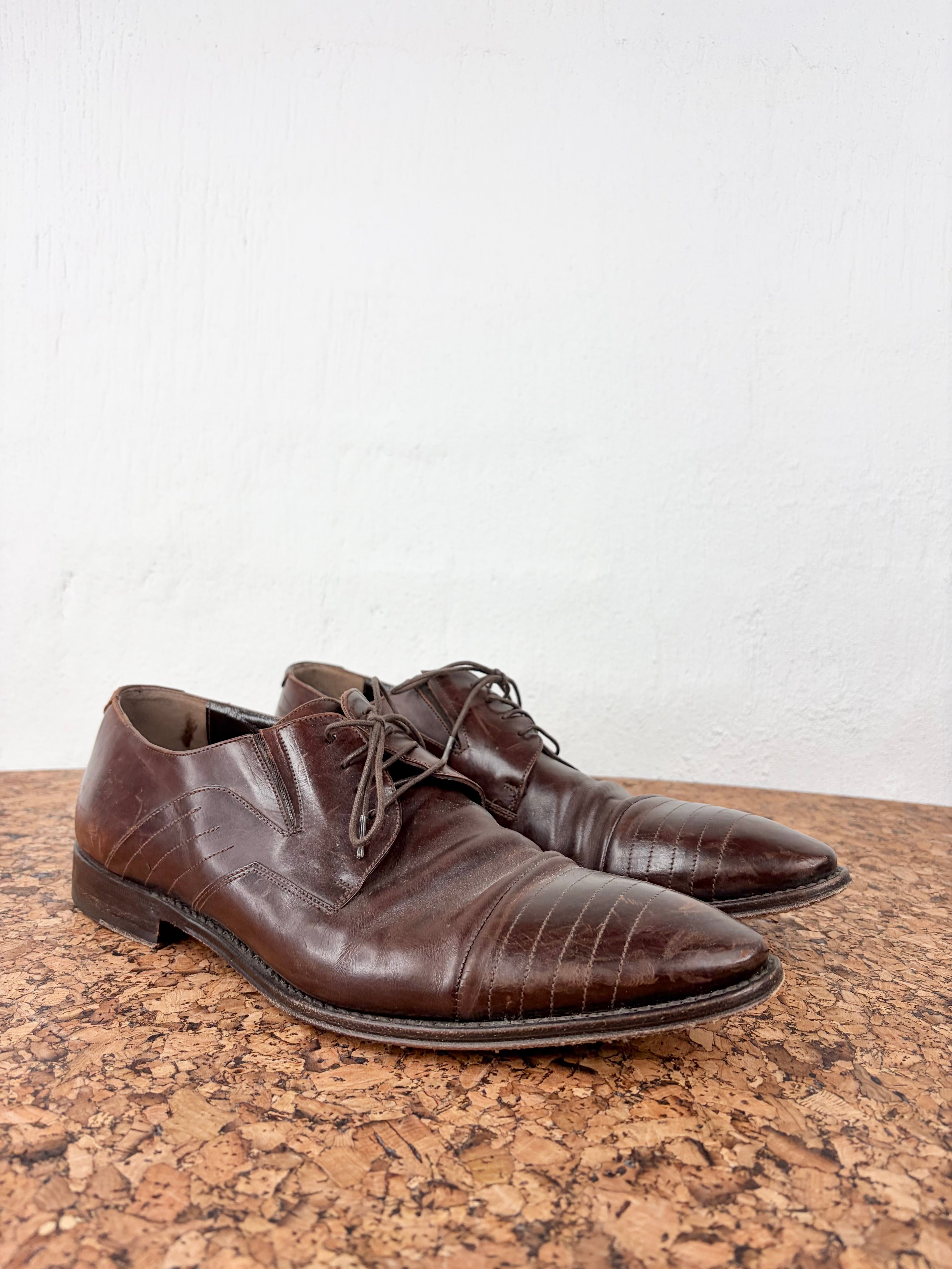 A pair of dark brown leather oxford shoes with a crocodile-embossed texture, displayed on a cork surface against a white wall.