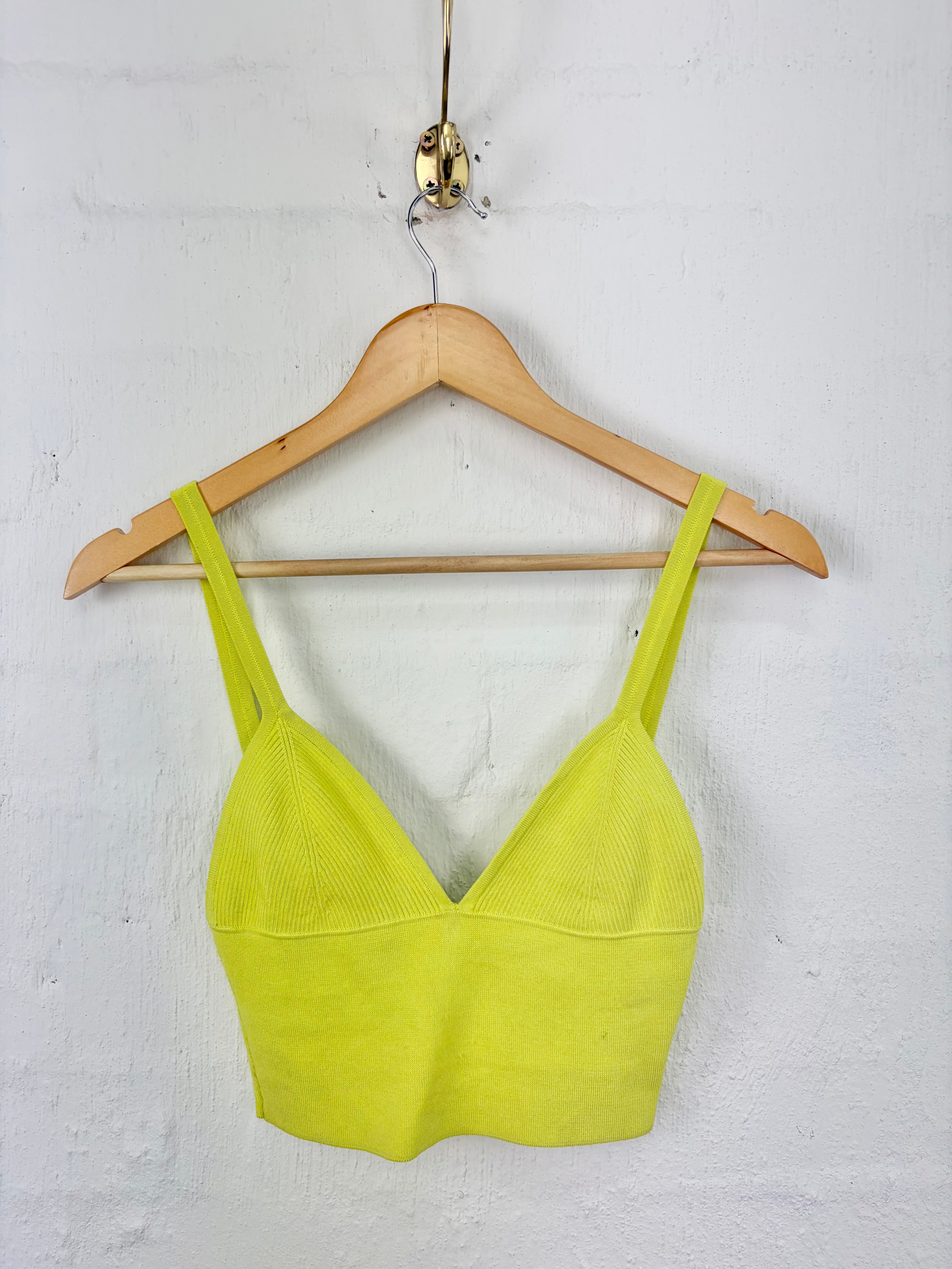 A bright neon yellow-green knit camisole top hanging on a wooden hanger against a white textured wall.