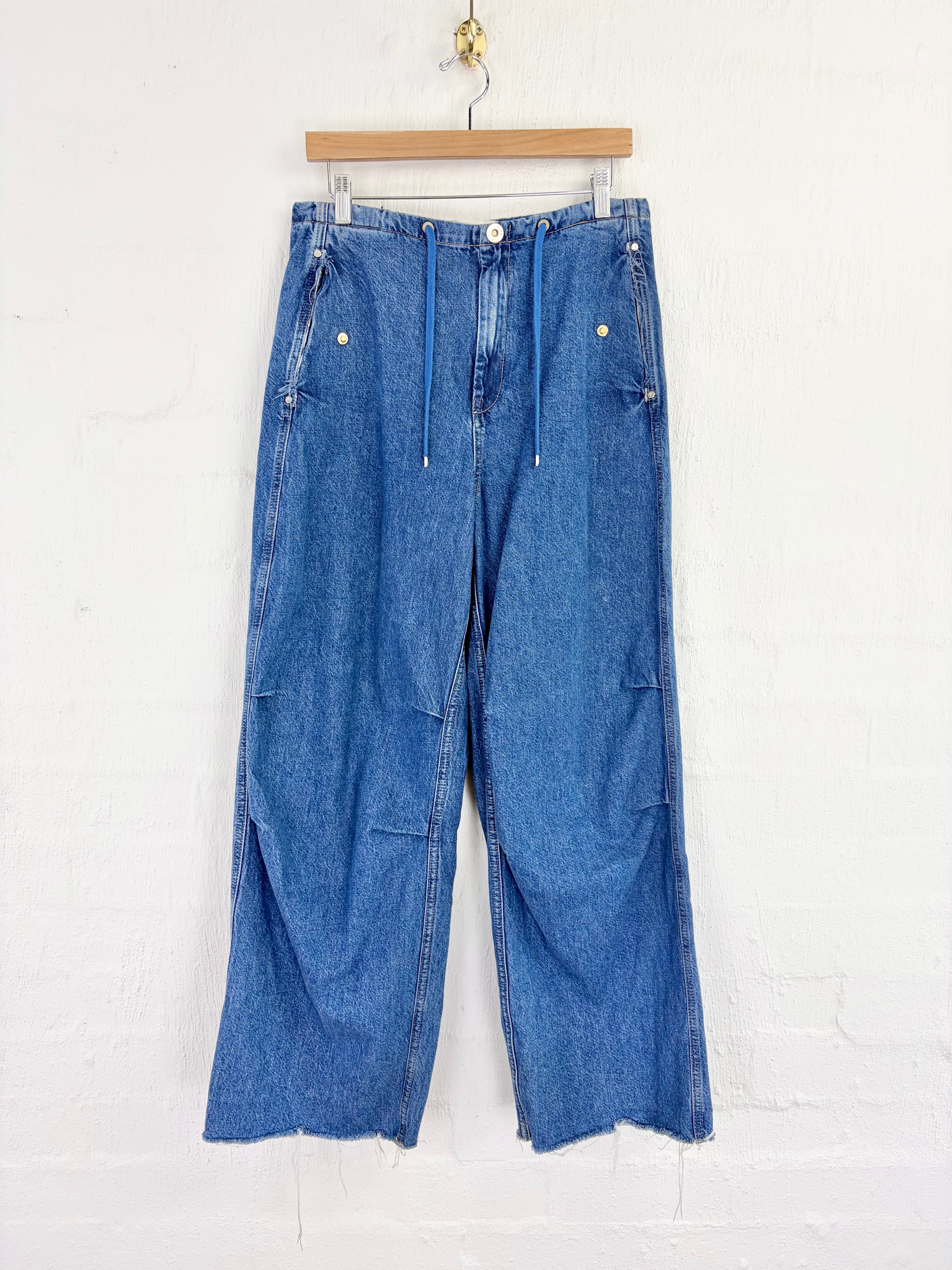 A pair of wide-leg blue denim pants with a drawstring waist, hanging from a wooden hanger against a white painted brick wall.