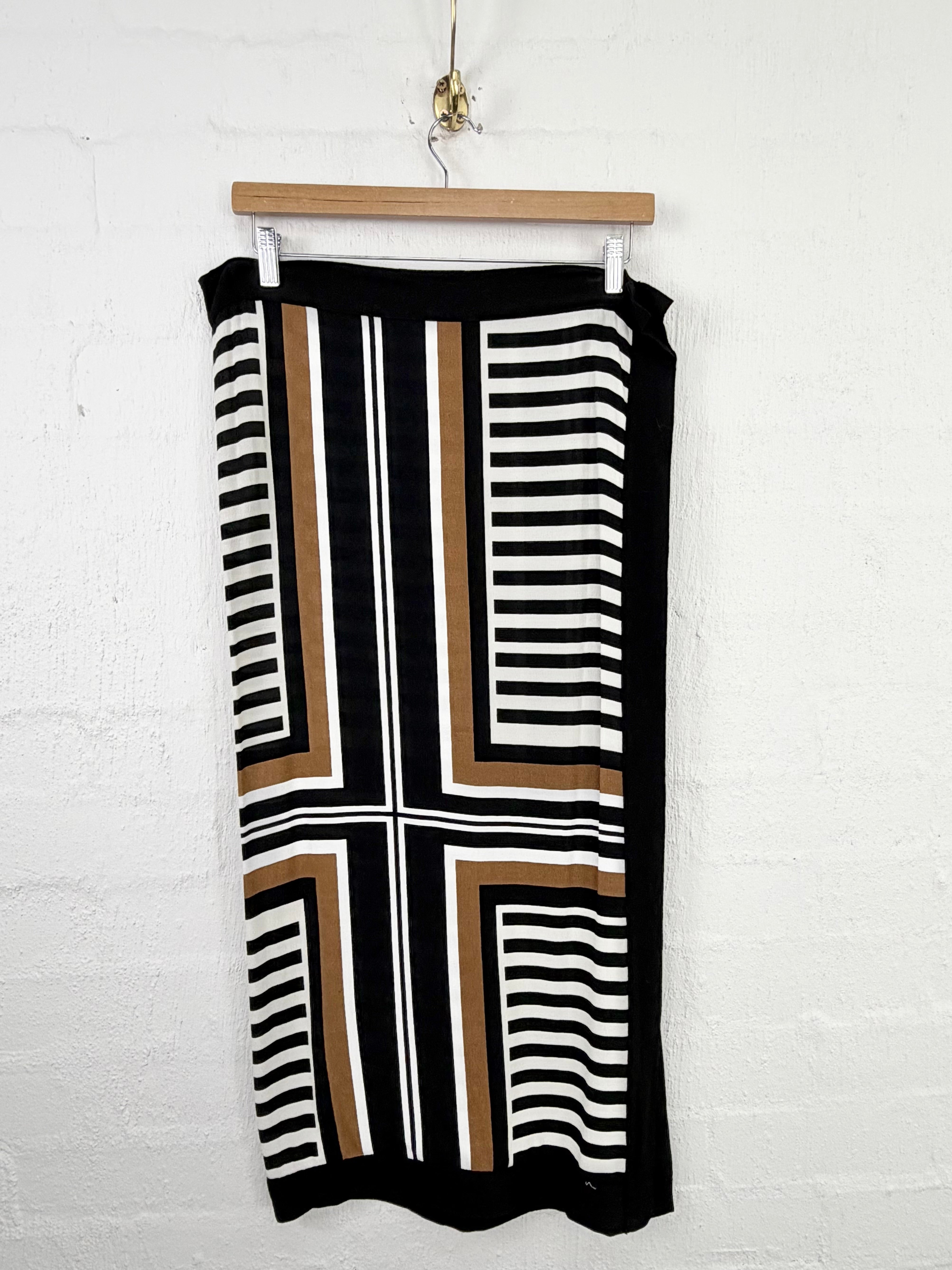 A sleeveless knit midi skirt with a geometric striped pattern in black, white, and brown, hanging from a wooden hanger against a white brick wall.