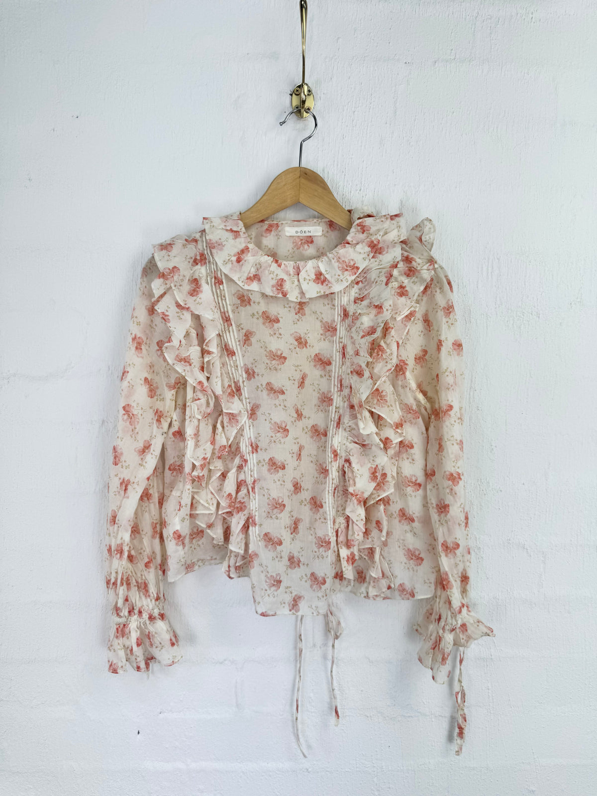A long-sleeve ruffled blouse with a small floral print, hanging on a wooden hanger against a white painted brick wall.