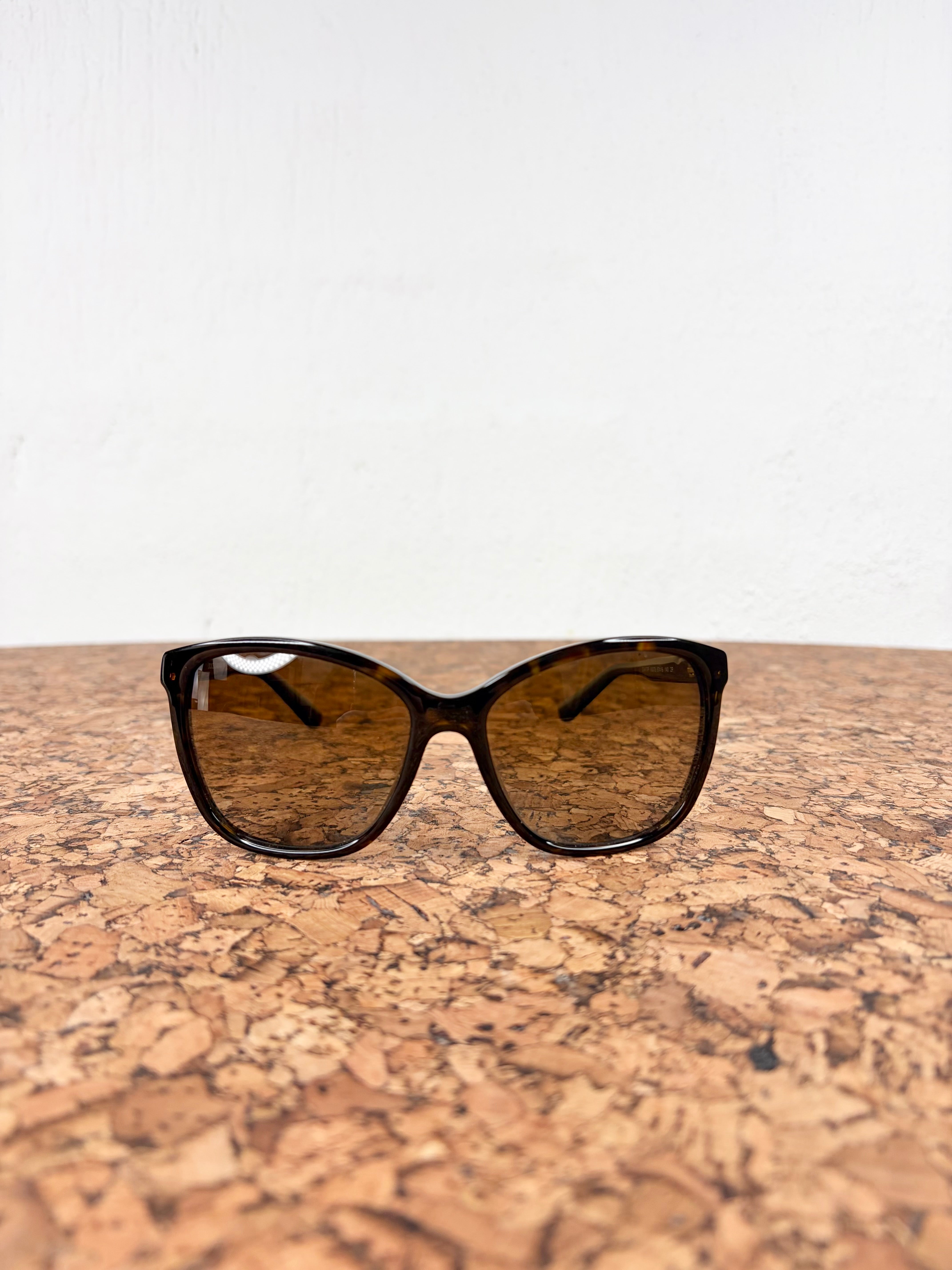 Cat-eye sunglasses with a dark tortoiseshell frame and brown tinted lenses, resting on a cork surface against a white wall.