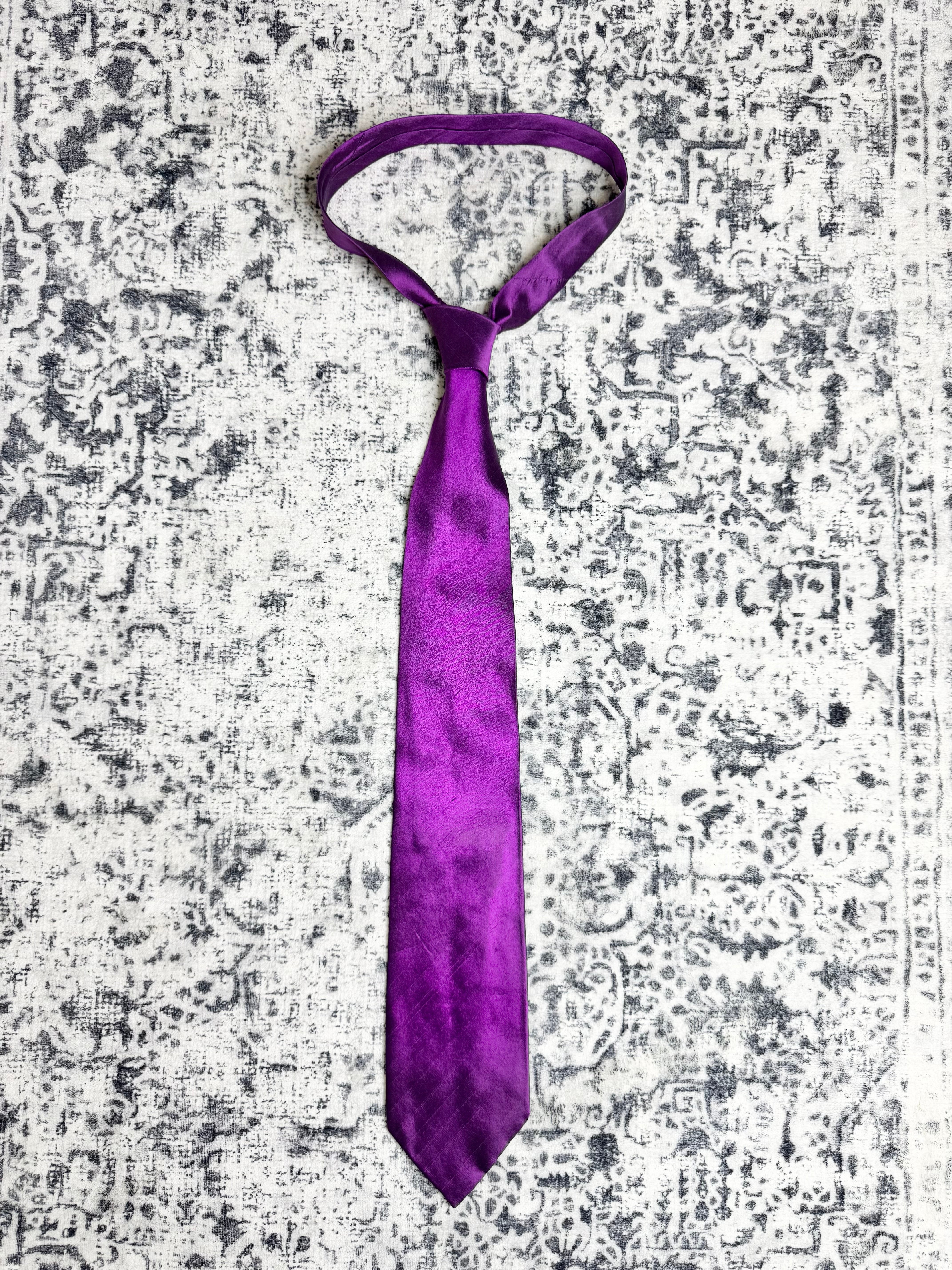 A purple satin necktie with a subtle sheen and textured pattern, laid flat on a distressed gray and white patterned rug.