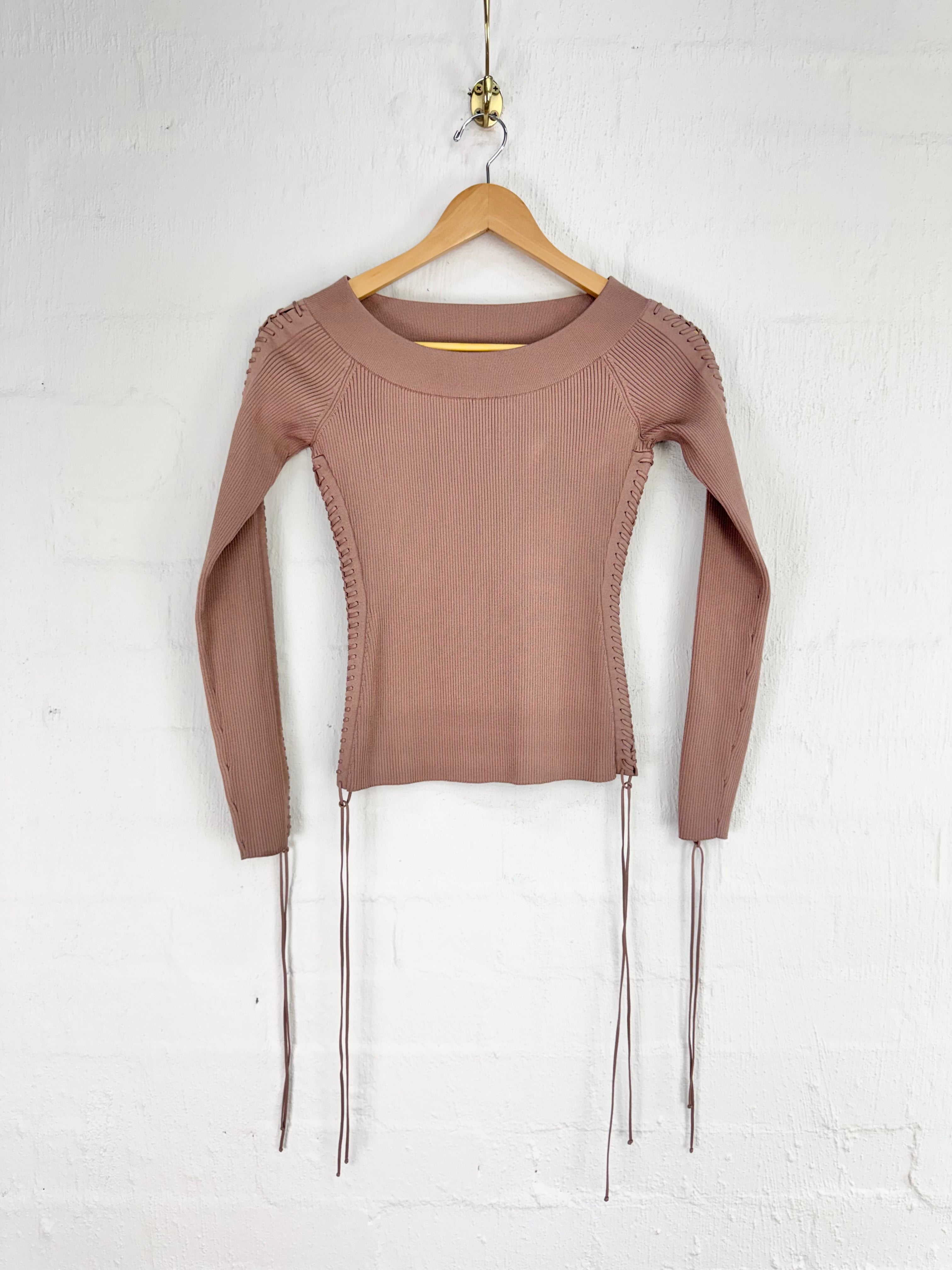 A long-sleeve knit top in a muted taupe-brown color, hanging on a wooden hanger against a white painted brick wall. The top features decorative braided detailing along the shoulders and sides, with thin dangling ties hanging from the hem.
