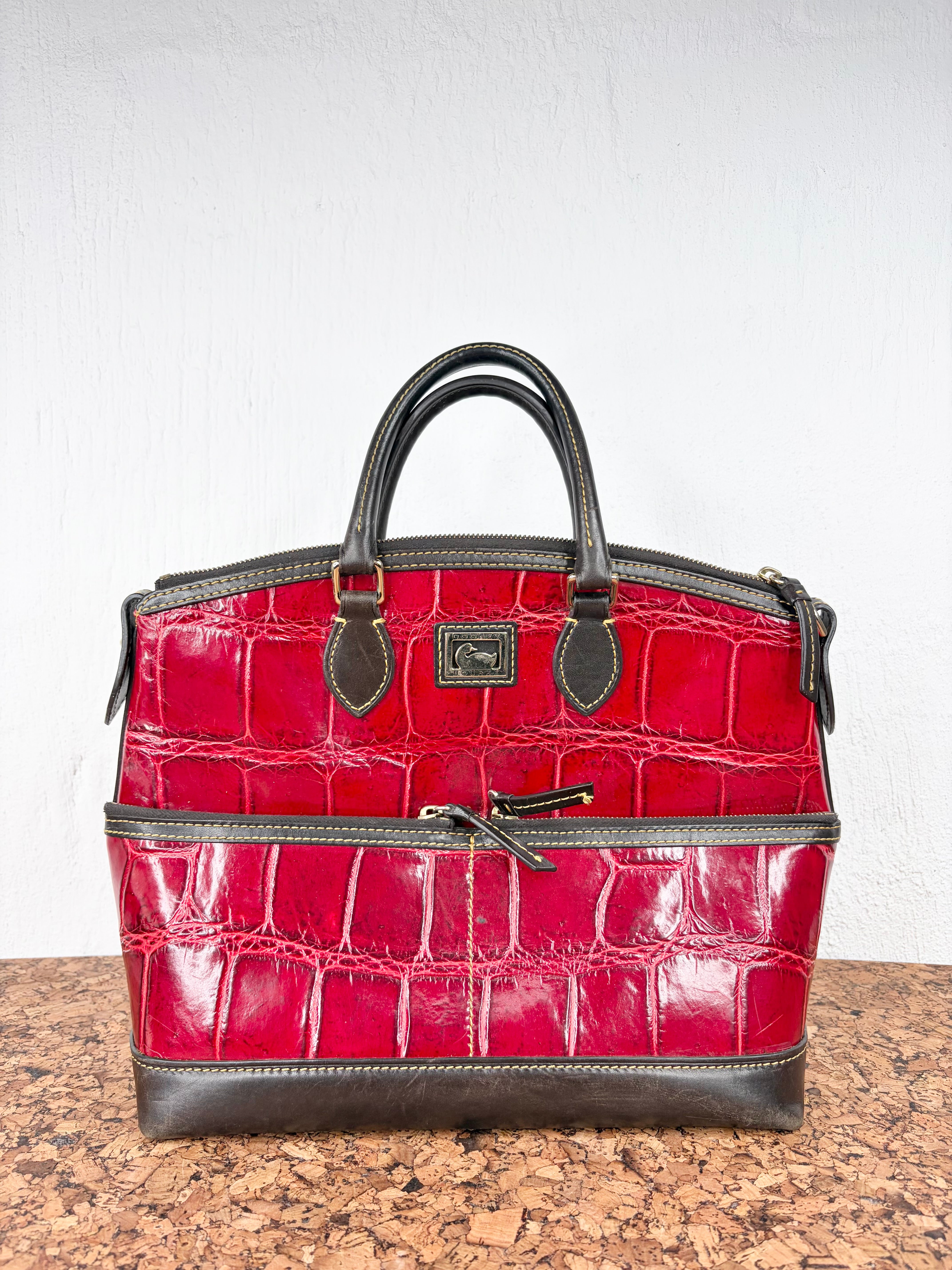 A structured red crocodile-embossed leather handbag with dark brown leather trim, double top handles, and a front zippered pocket, displayed on a speckled brown countertop against a white wall.