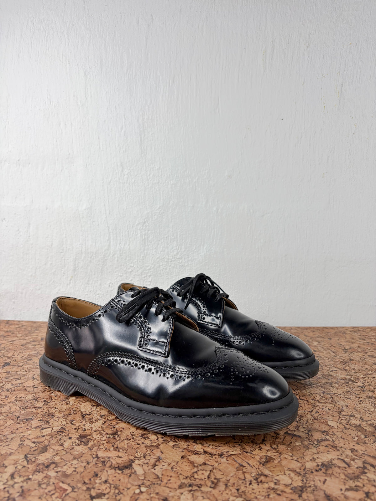 A pair of glossy black leather wingtip oxford shoes with brogue detailing, displayed on a brown speckled countertop against a white textured wall.