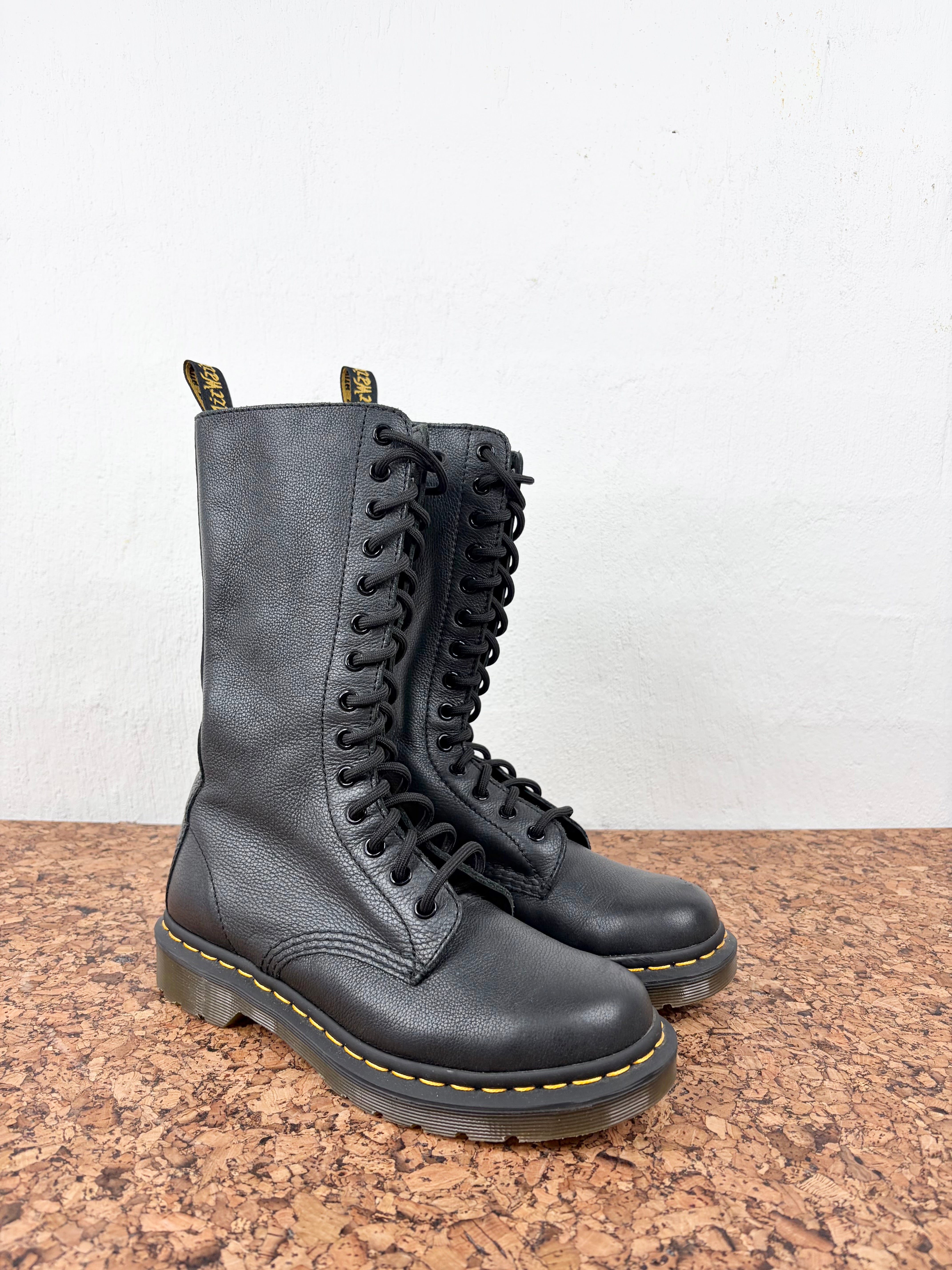 A pair of black leather Dr. Martens 16-inch lace-up combat boots with yellow stitching and black soles, displayed on a brown cork surface against a white wall.
