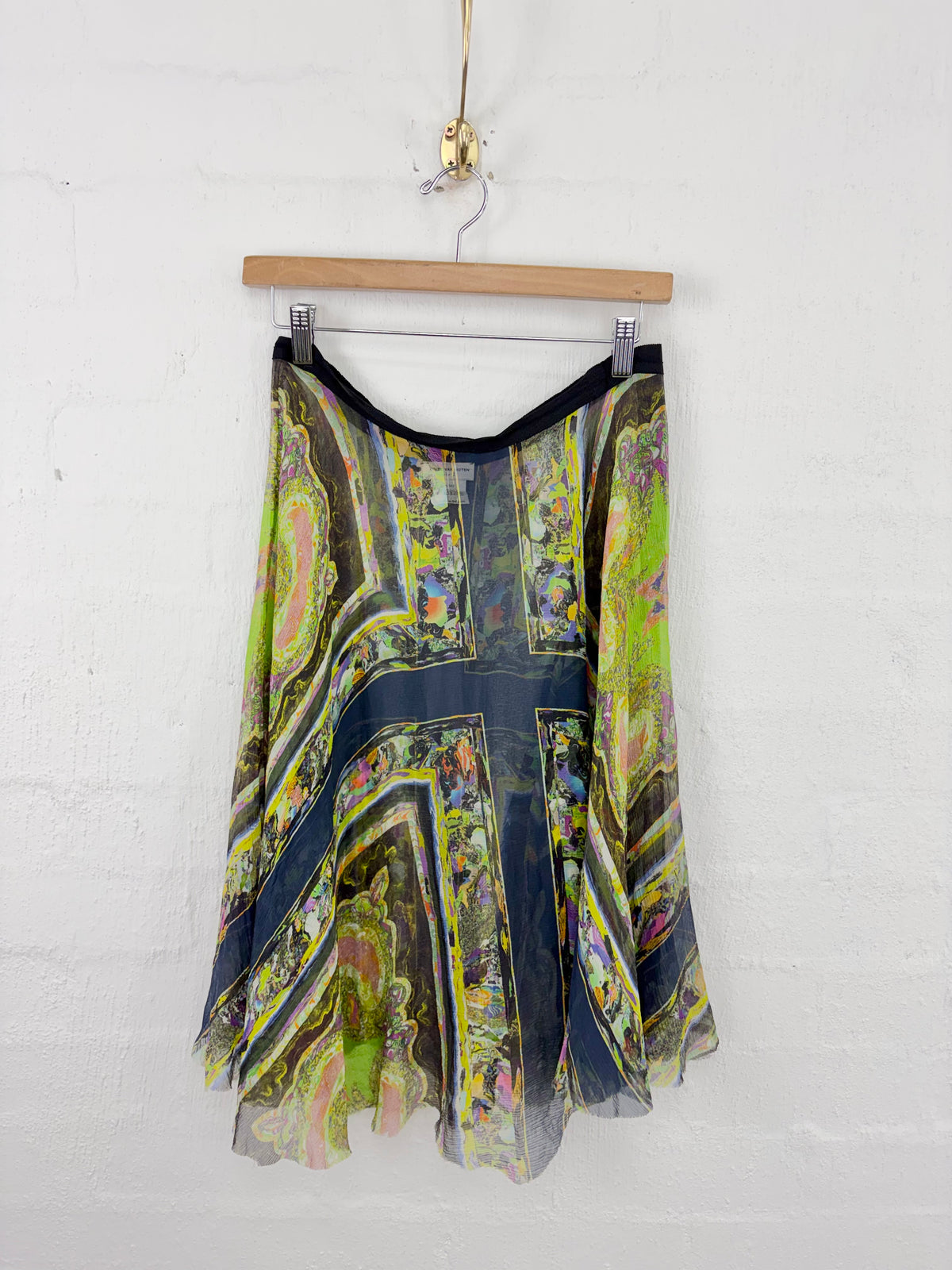 A sheer, lightweight A-line skirt with a vibrant abstract print in green, yellow, black, and blue, hanging from a wooden hanger against a white painted brick wall.