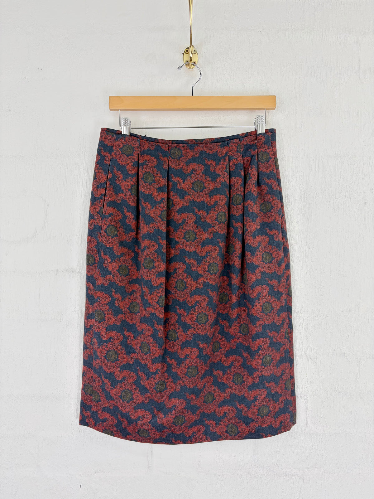 A pleated A-line skirt with an intricate red and navy blue floral pattern, hanging from a wooden hanger against a white painted brick wall.