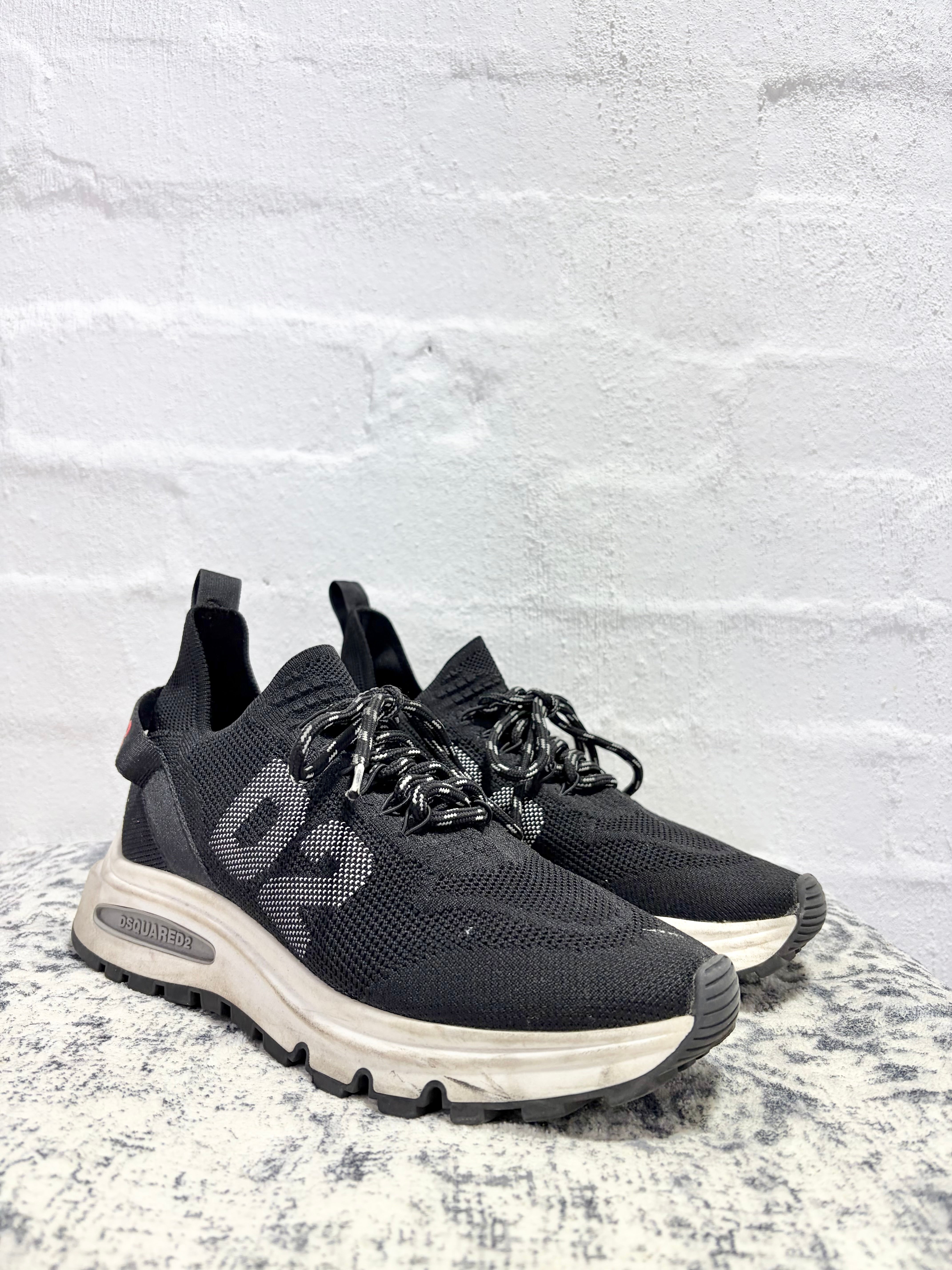 A pair of black athletic sneakers with chunky off-white midsoles, displayed on a speckled stone surface against a white brick wall background.