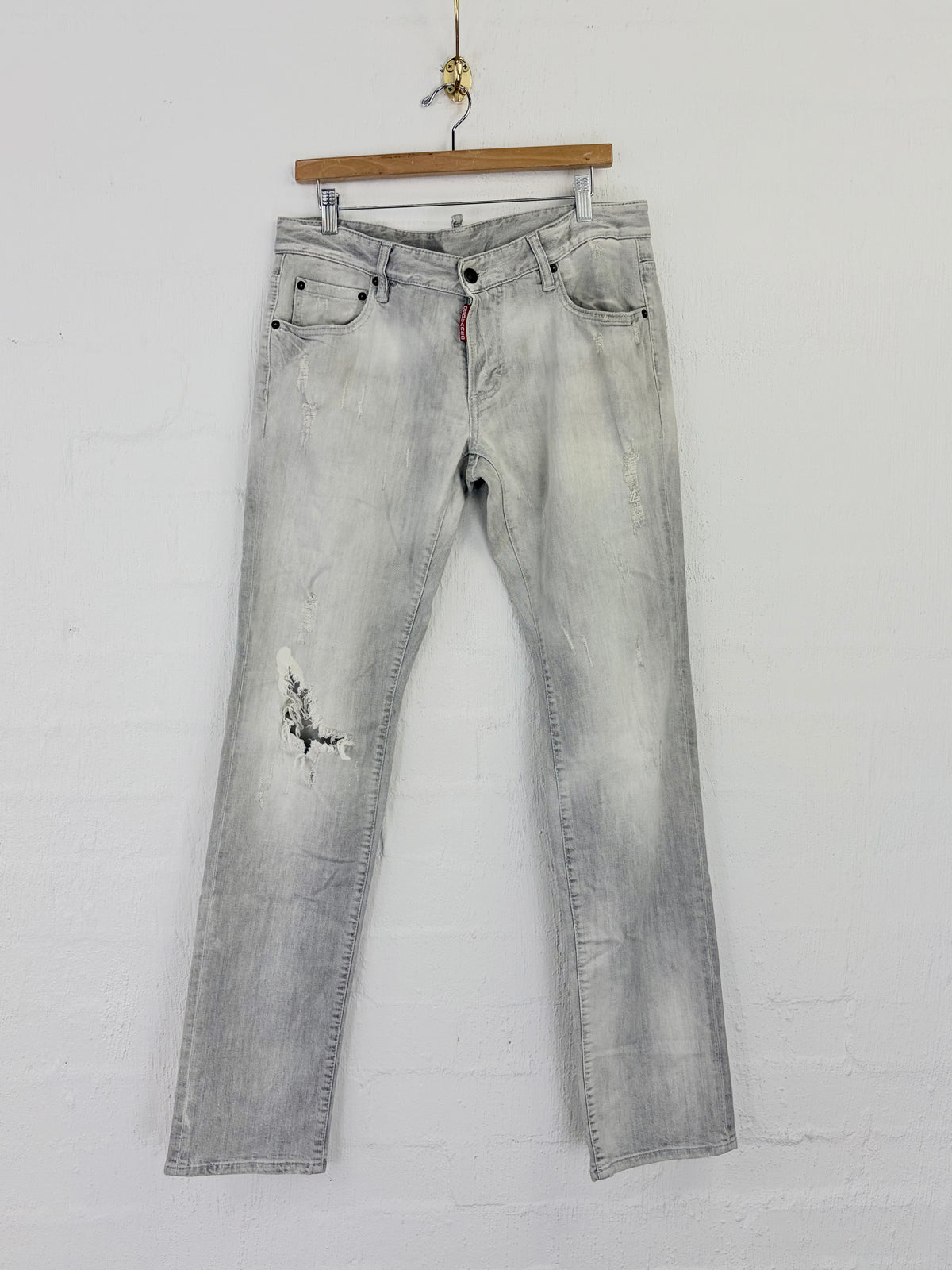Light gray washed denim jeans with distressed details on the left thigh, hanging from a wooden hanger against a white brick wall.