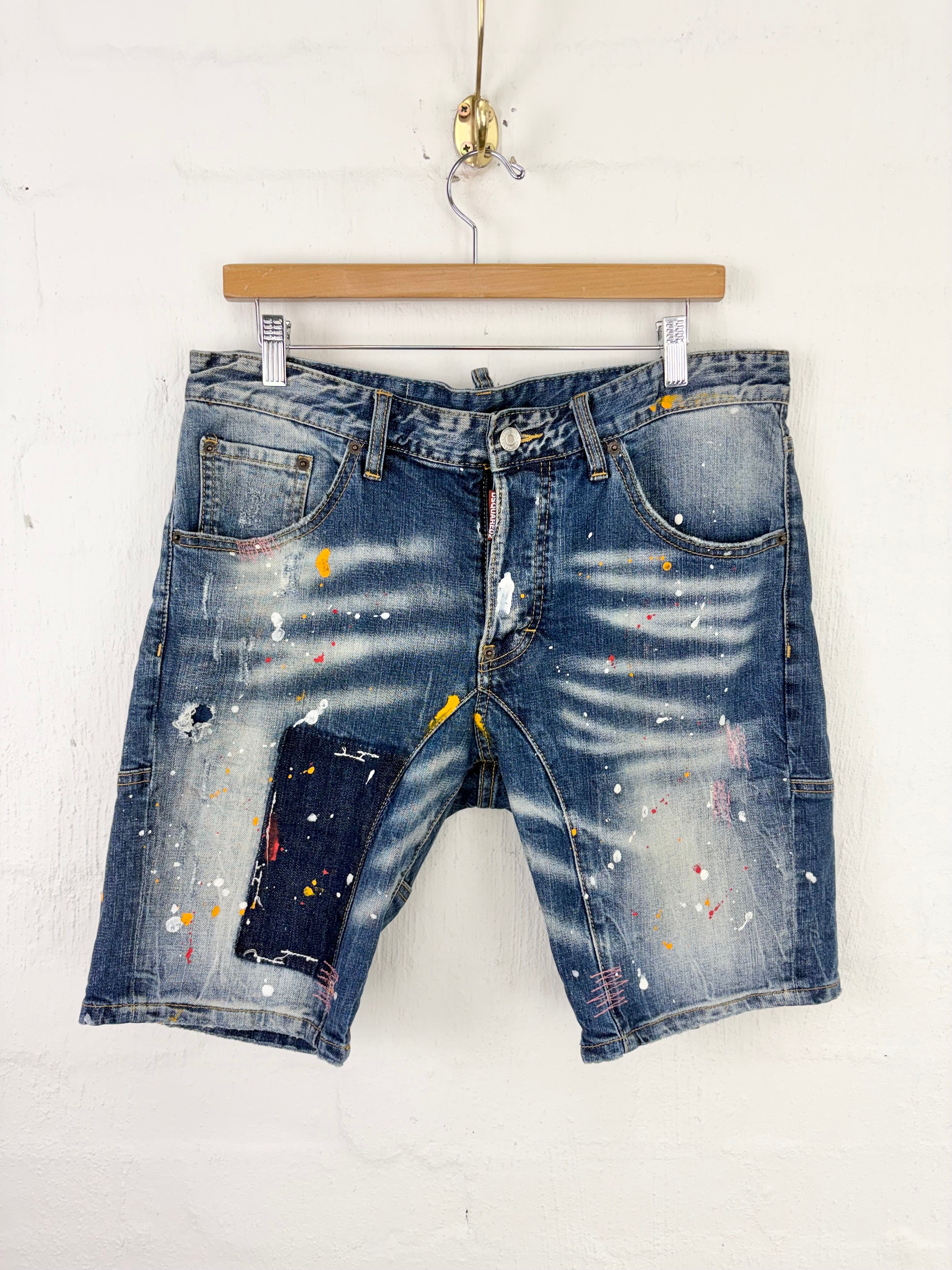 Distressed blue denim shorts with paint splatter details, hanging from a wooden hanger against a white brick wall.