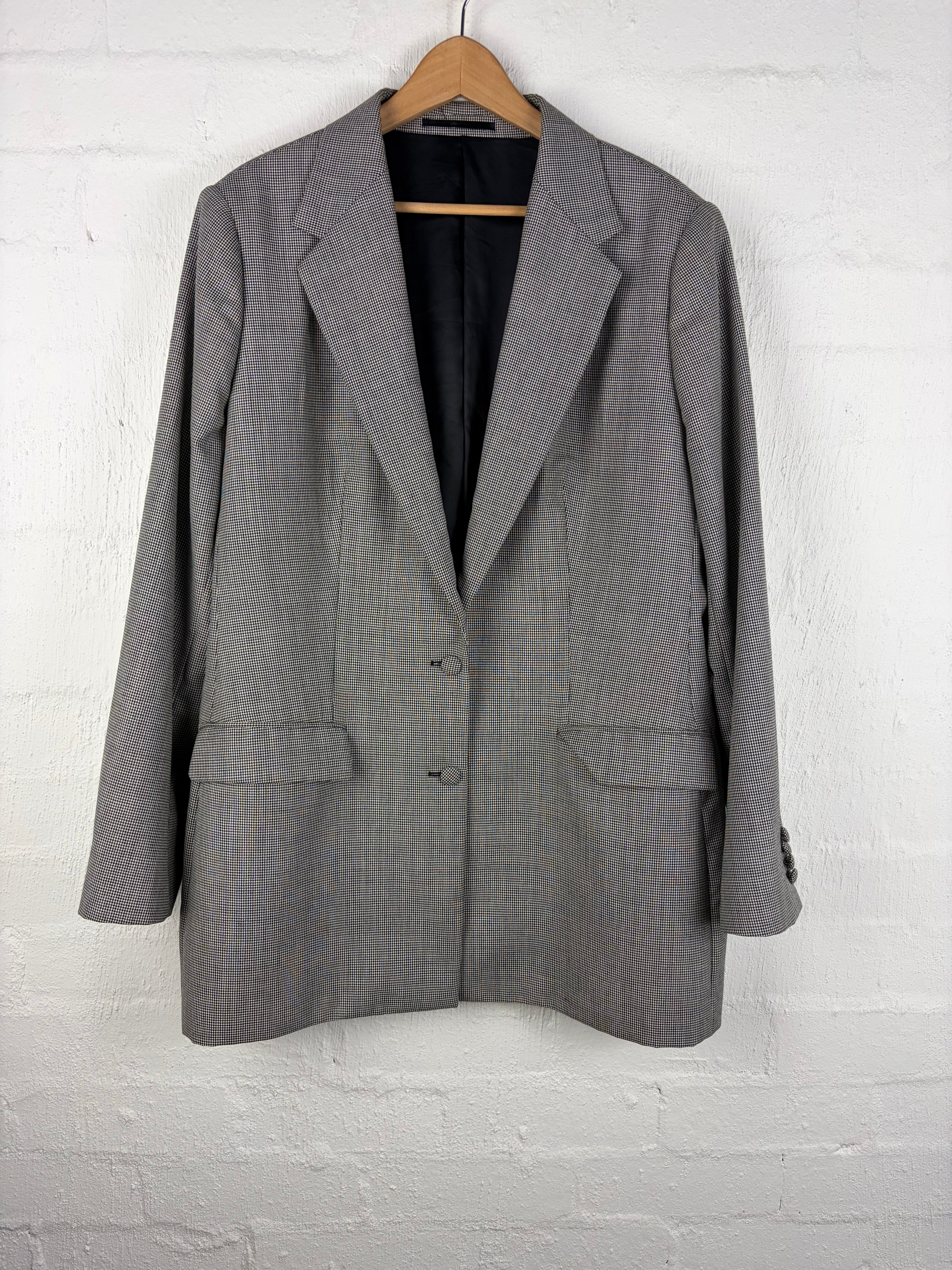 A gray micro-check blazer jacket hanging on a wooden hanger against a white painted brick wall.