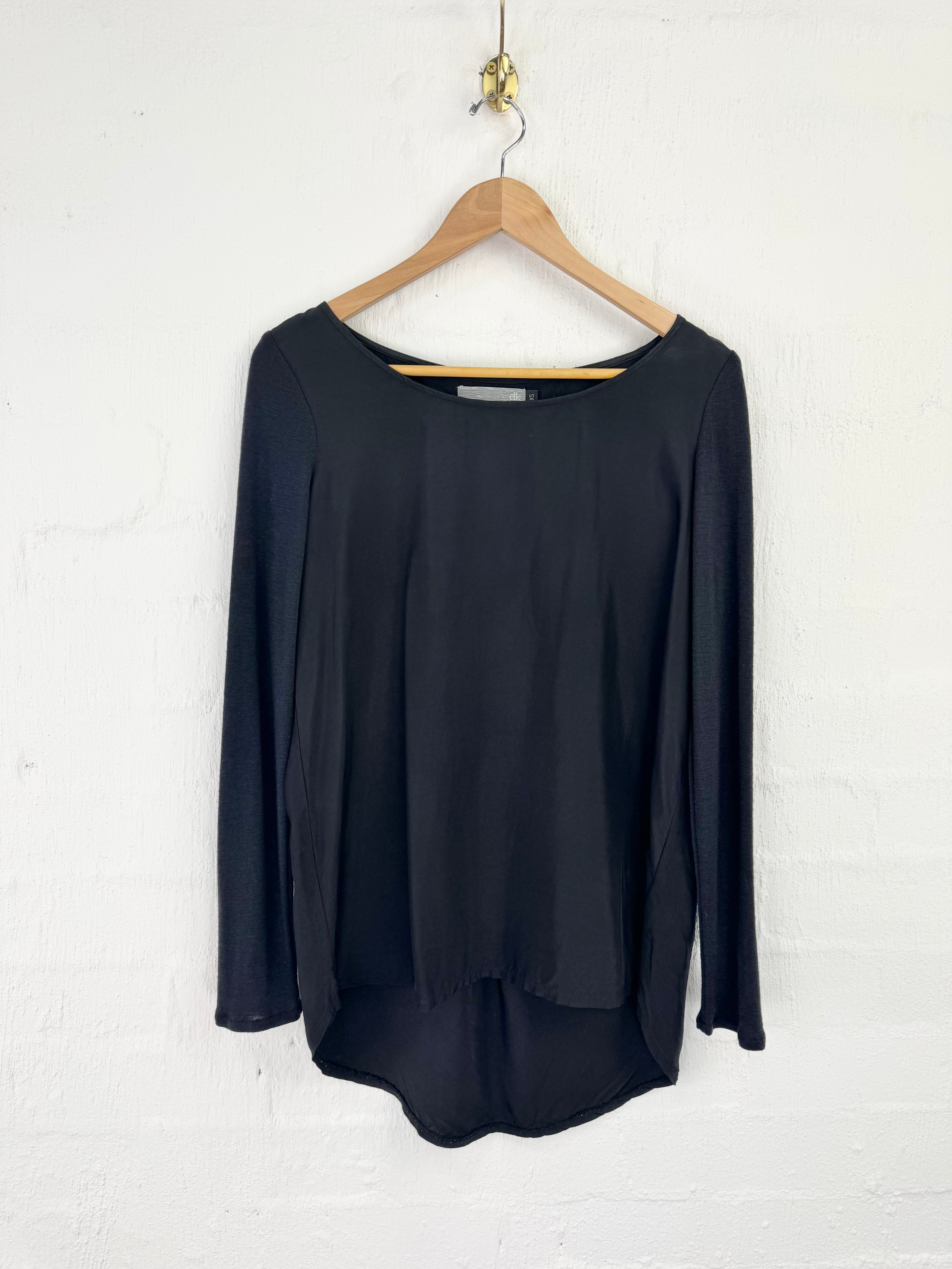 A black long-sleeve top with a wide boat neckline, hanging on a wooden hanger against a white painted brick wall.