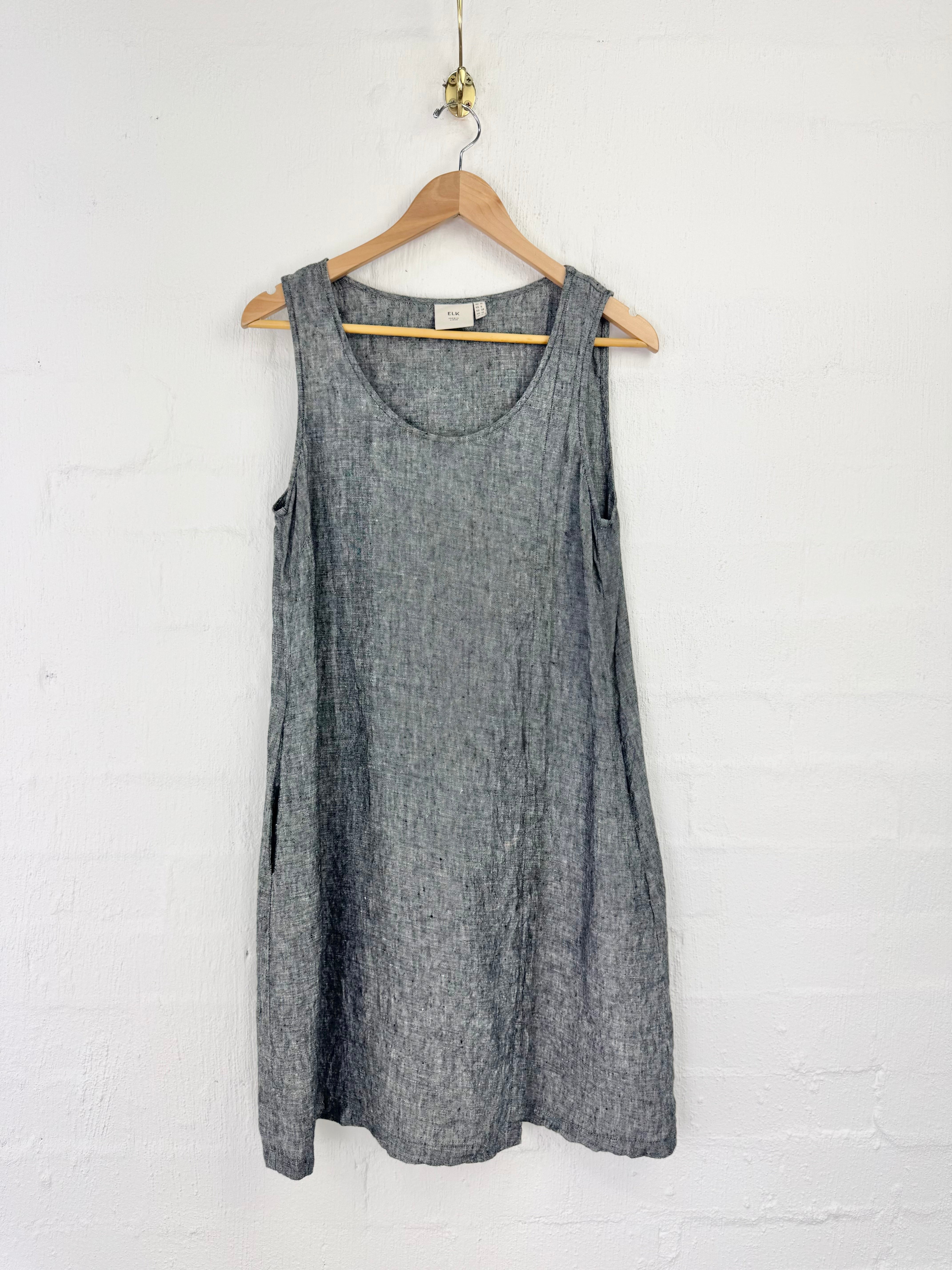 A sleeveless gray linen dress hanging on a wooden hanger against a white painted brick wall.