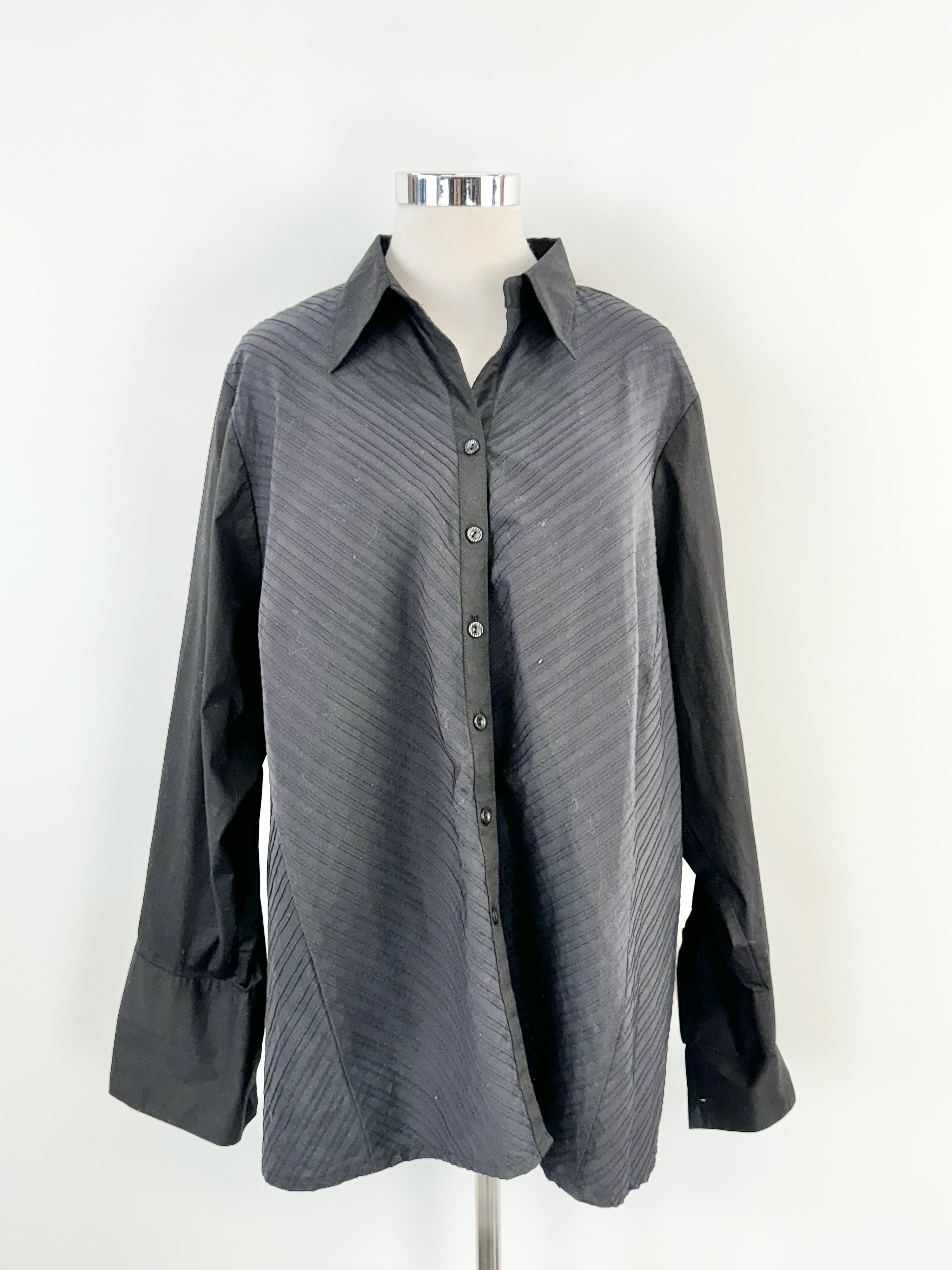 An oversized black blouse with a sharp, crisp collar and pintuck details on the body. The sleeves are smooth and black, ending with a long French cuff.