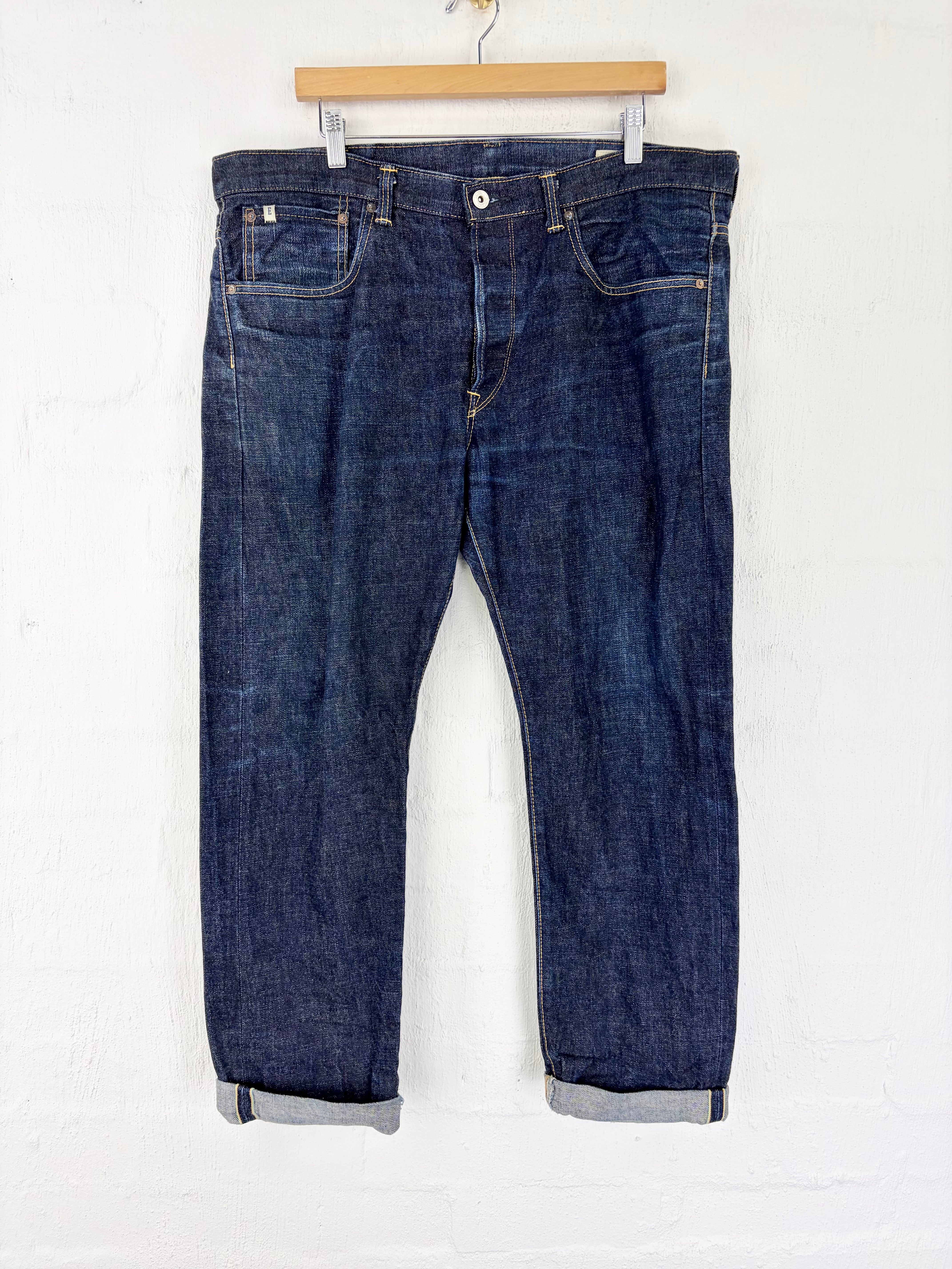 A pair of dark blue denim jeans with rolled cuffs, hanging from a wooden hanger against a white brick wall.