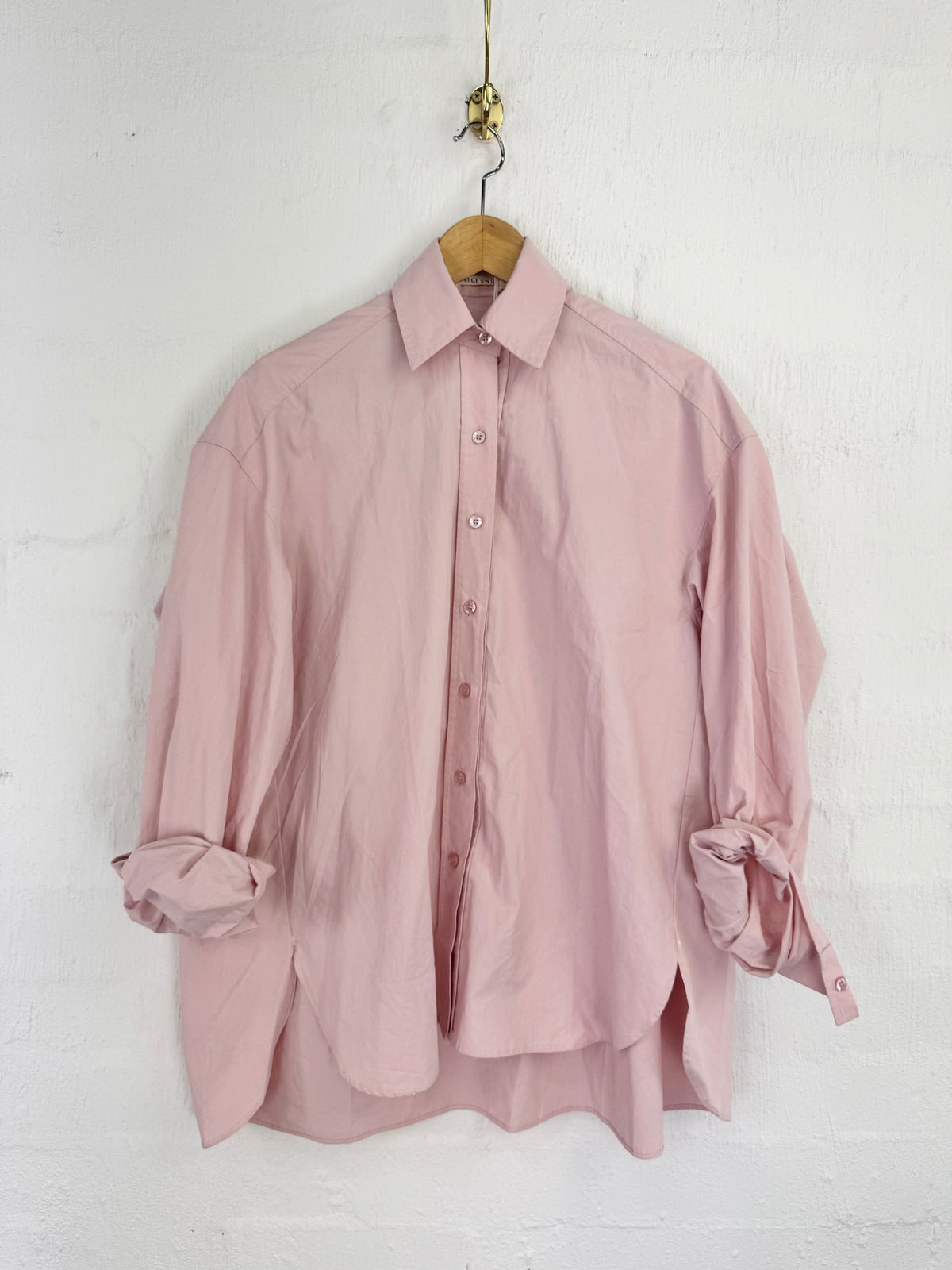 A dusty pink button-up shirt with rolled-up sleeves hanging on a wooden hanger against a white textured wall.