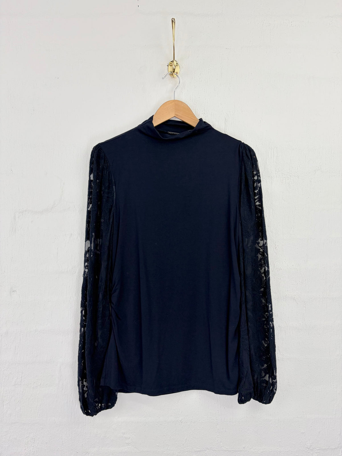 A dark navy blue long-sleeve mock neck top with sheer patterned sleeves, hanging on a wooden hanger against a white painted brick wall.